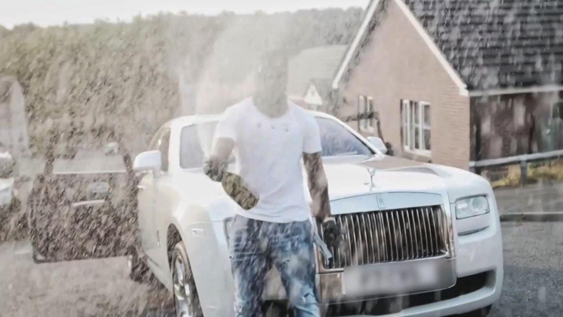 A man in a white t-shirt sprays champagne in front of a luxury car