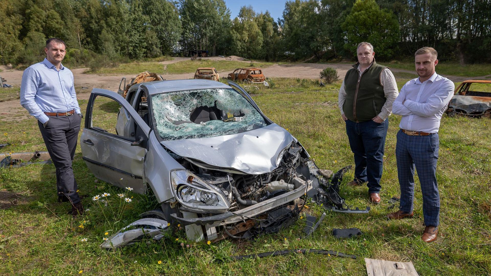 Three men stand around a smashed up small car in the former Torvean Quarry. There are burnt out, rusting cars behind them. The area of the quarry is surrounded by trees.