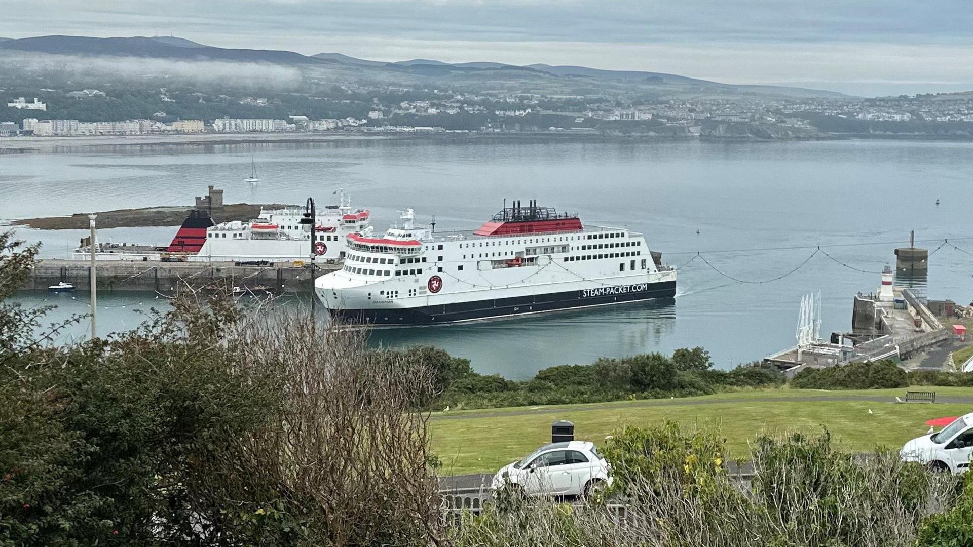 The Manxman ferry, which is red, white and black, entering Douglas Harbour. The Ben-my-Chree is moored at the other side of the pier behind it and there cars parked on the headland overlooking the harbour.