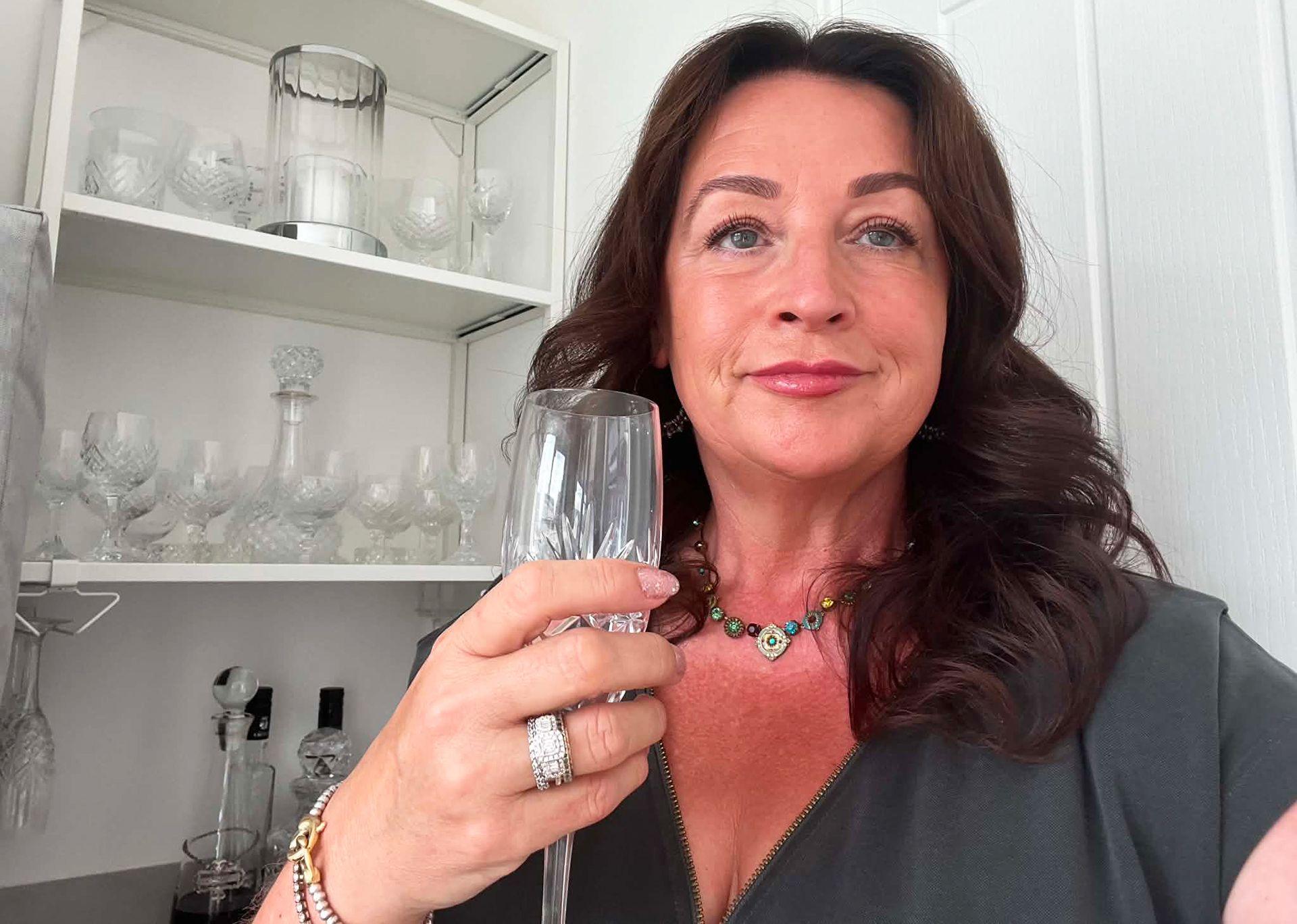 Woman with long dark hair and thoughtful expression is holding a champagne glass. A large ring is visible on the fourth finger of her hand holding the glass.