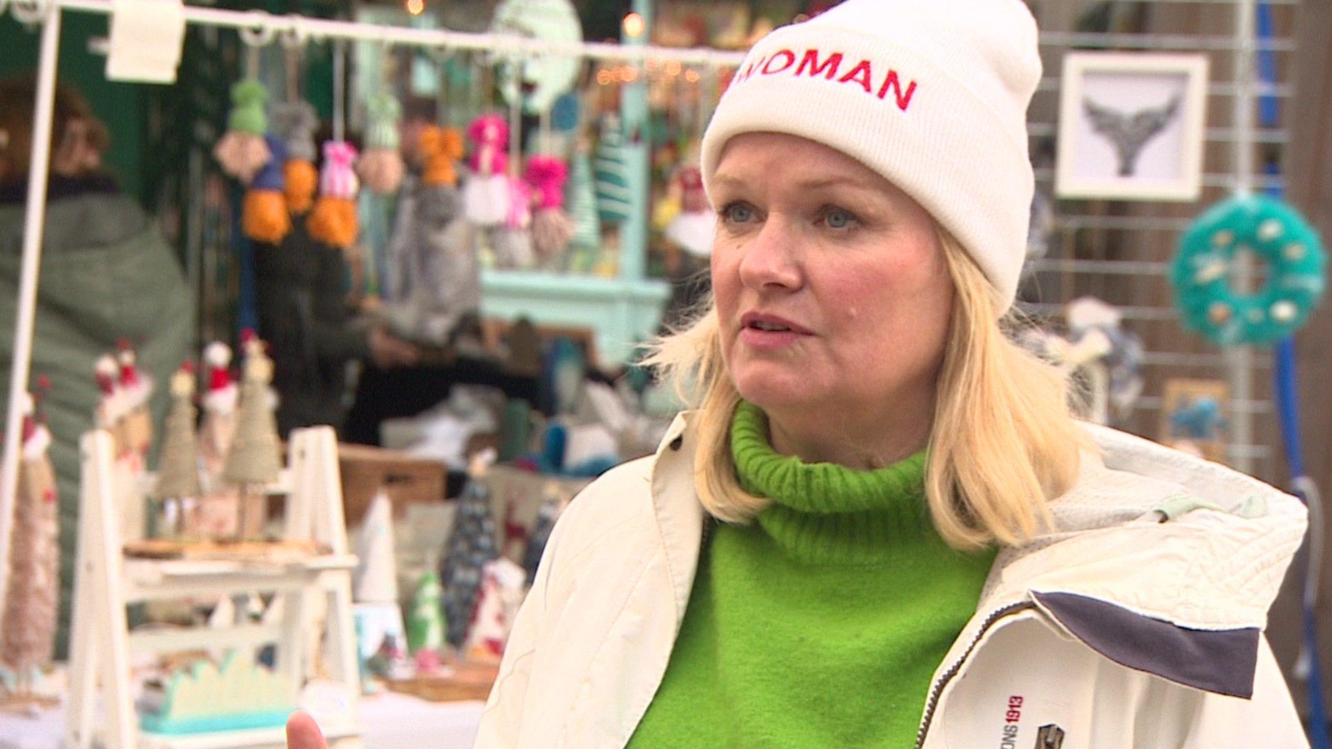 A woman with blonde hair wearing a white beanie hat with 'Woman' written in red letters on front, wearing a green roll neck sweater and a cream jacket in front of a market stall