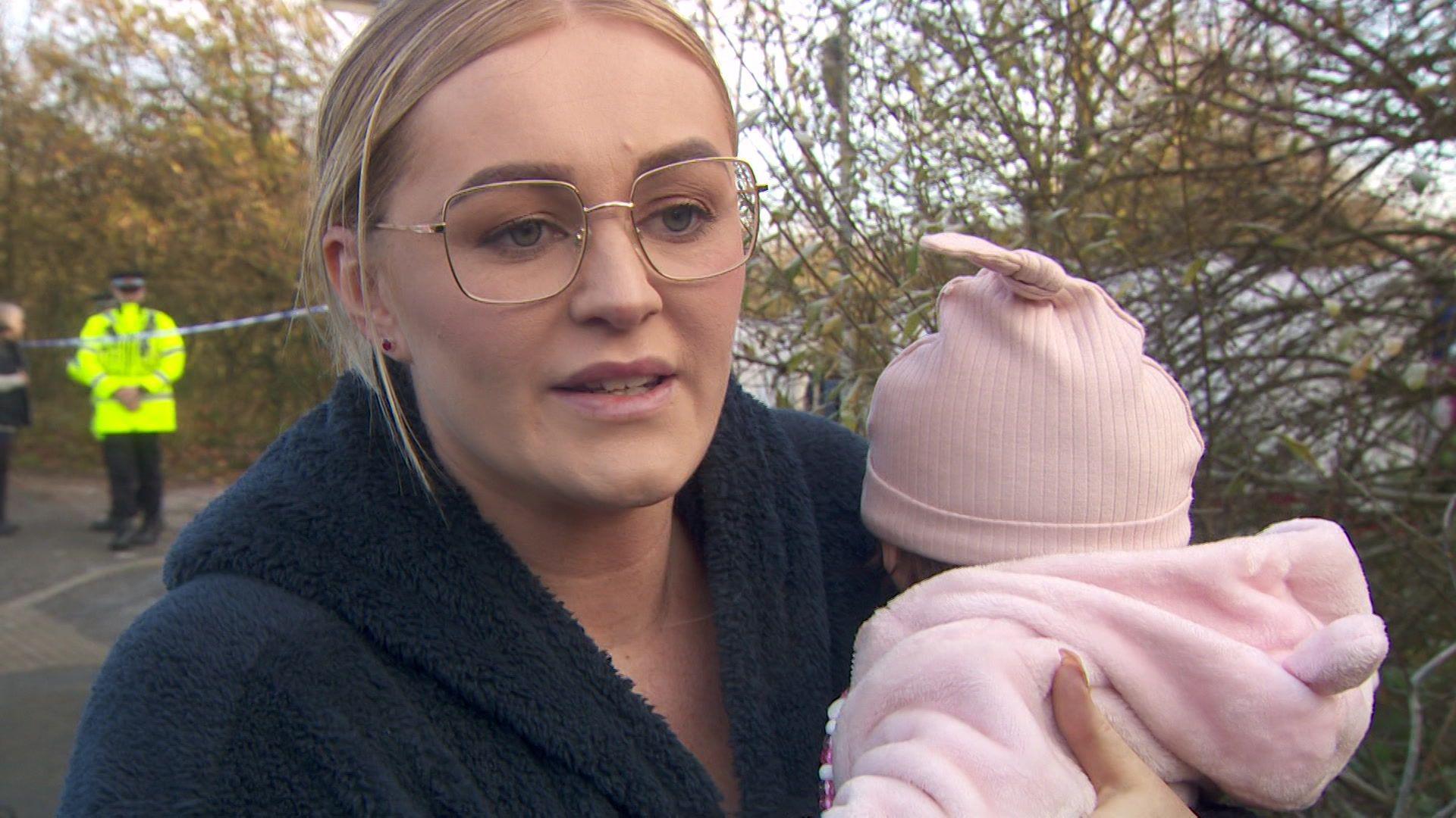 Dawn Thompson, with blonde, tied back hair and wearing glasses and a black coat, holds her baby who is wearing a pink hat and pink fluffy coat.