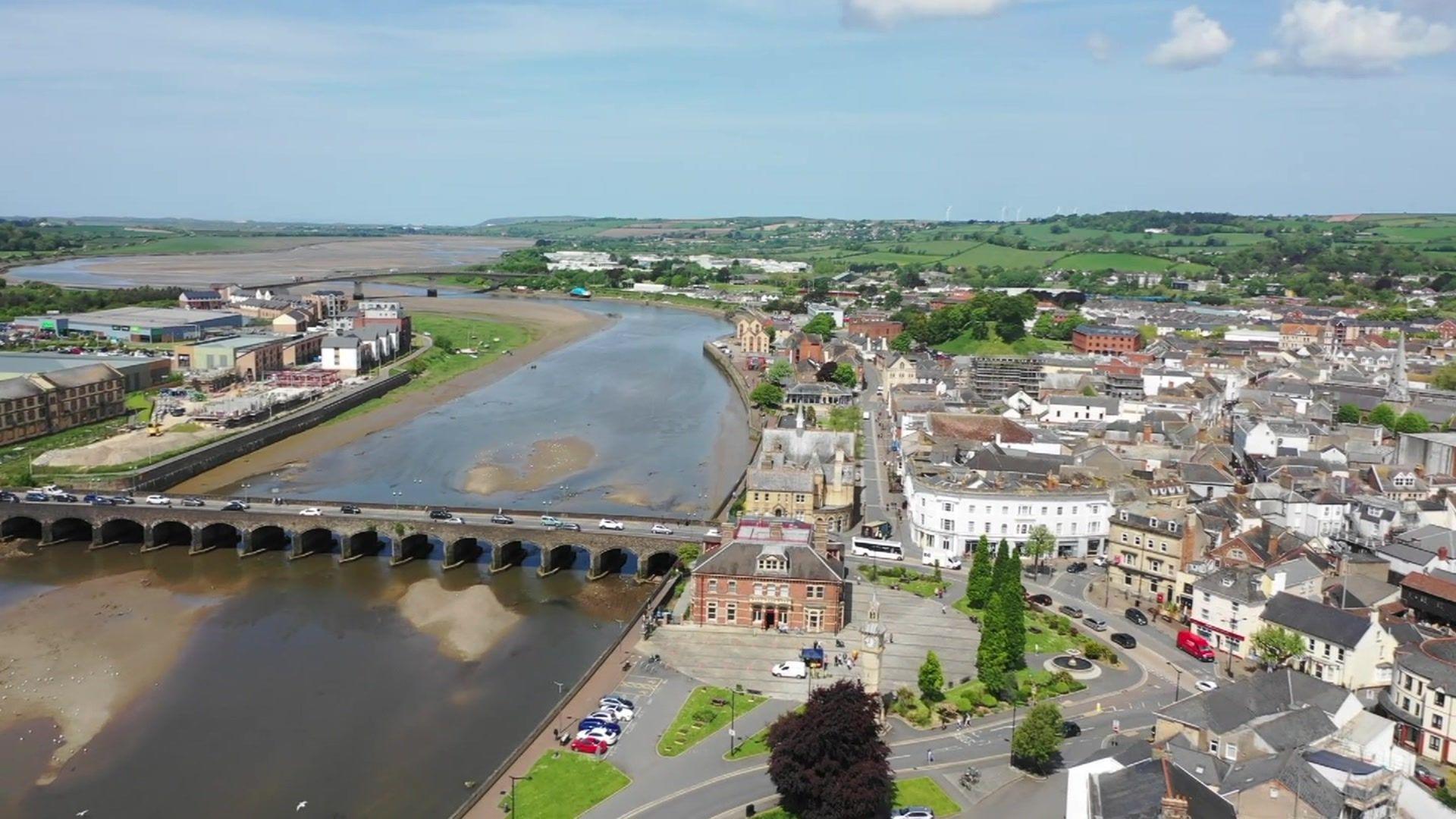 Aerial view of the River Taw. There is land to the right of the river with vehicles, a main road and numerous buildings. On the left a factory-type building can be seen in the distance. A bridge can be seen with cars travelling on it.