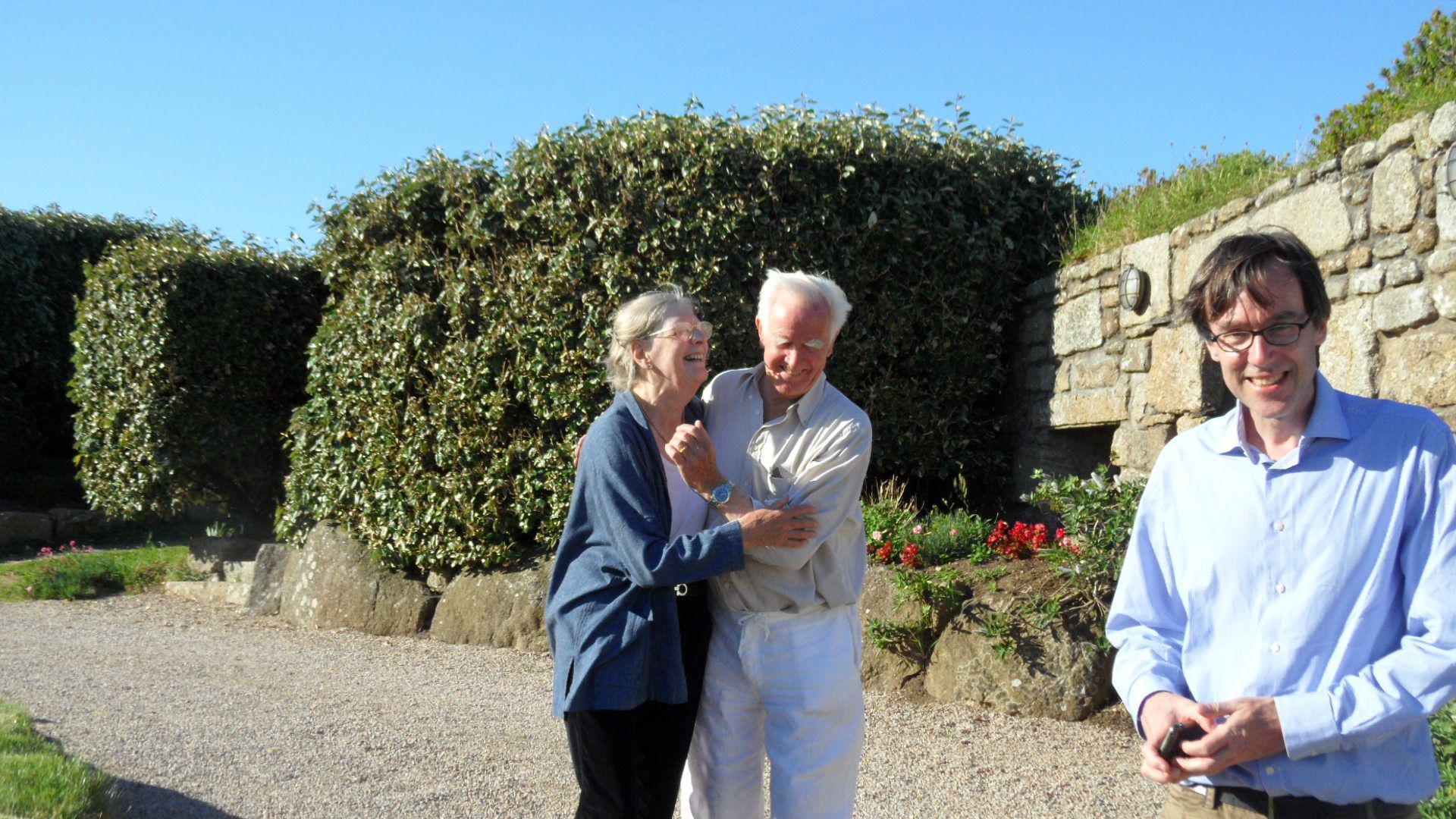 A man and a woman laughing with their arms round each other, standing on a gravel driveway with a wall and a large bush behind them. A man stands on the right hand side, laughing and looking at the camera. It's a sunny day and the sky is blue.