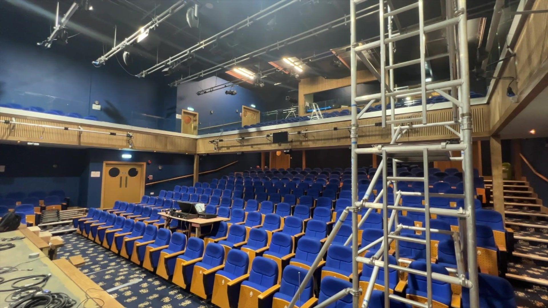 Around 10 rows of blue audience seats inside the theatre, with a scaffolding on the right hand side.