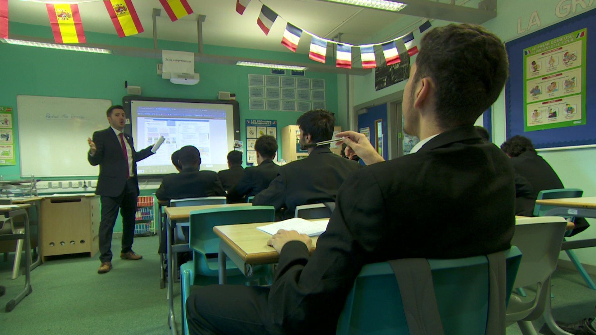 Pupil are sitting at desks in a classroom facing the board and a teacher is standing at the front. 