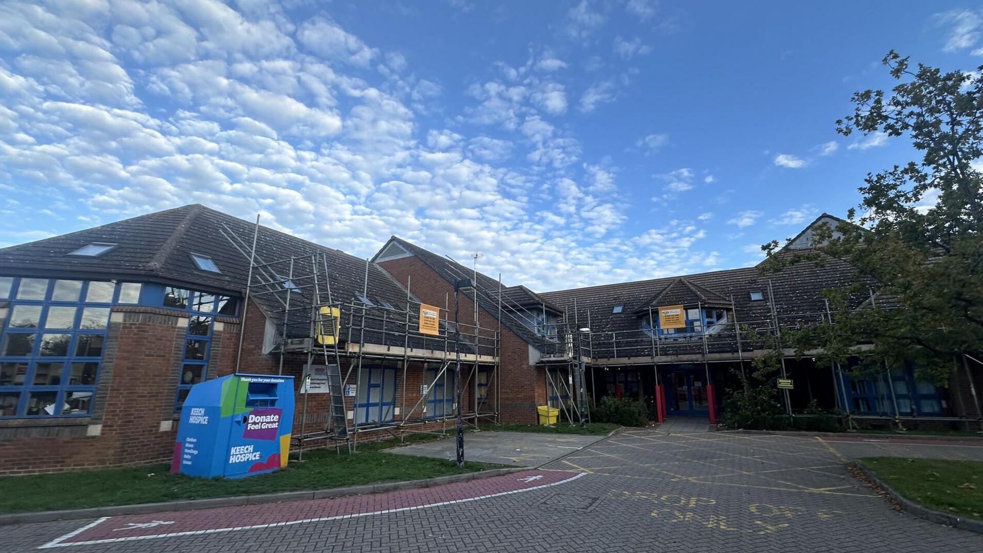 The front of Keech Hospice, which has scaffolding erected around the building. There is a large donations bin outside with Keech branding on. There is a yellow marked drop-off zone on the paved area outside.