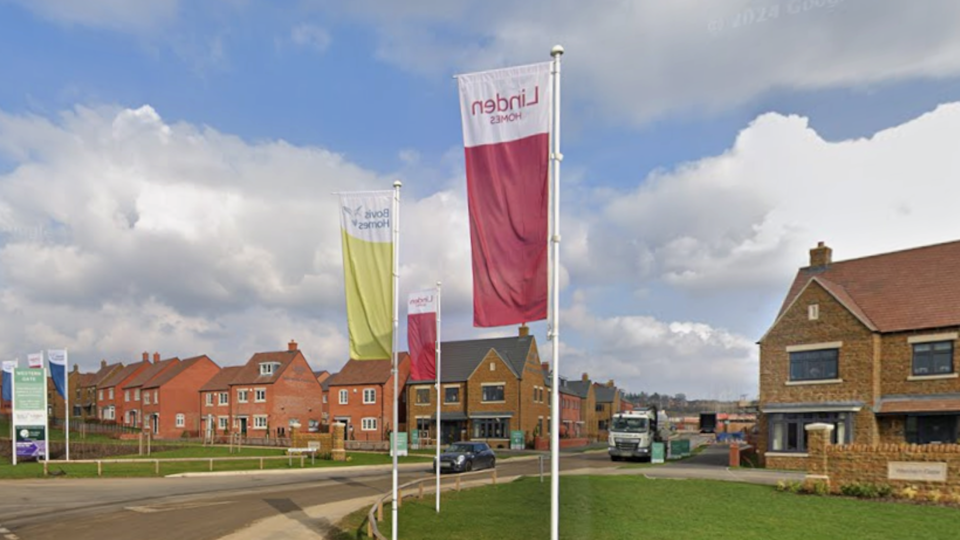 A new-build housing estate with red and brown brick houses set back from the road, with green space in front of them. Several white flagpoles are flying the flags of the homebuilders, including Linden Homes. There is a lorry and a car parked on the road.