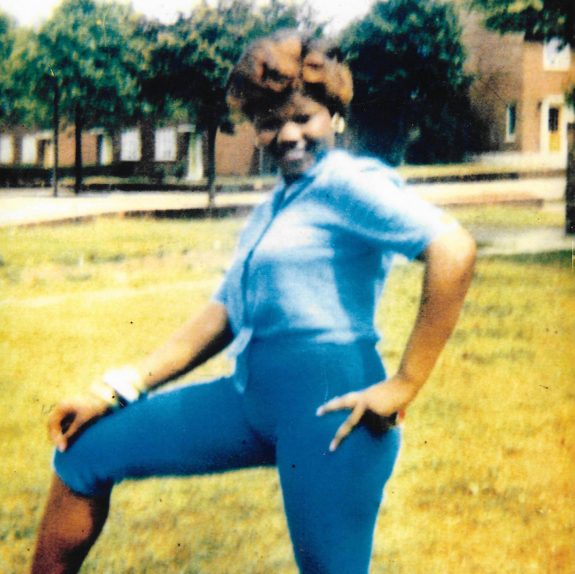 Olive Gordon in blue jeans and blue shirt is smiling broadly and posing for the camera with her left hand on her hip. She has short, spiky hair. She is on a lawn with modern houses behind her.