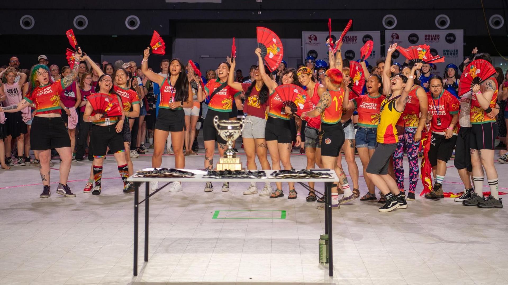 Team Chinese Nations celebrating the win of the Spirit Award at the Roller Derby World Cup. The members are cheering and waving folding fans. There is cup on a table in front of them.