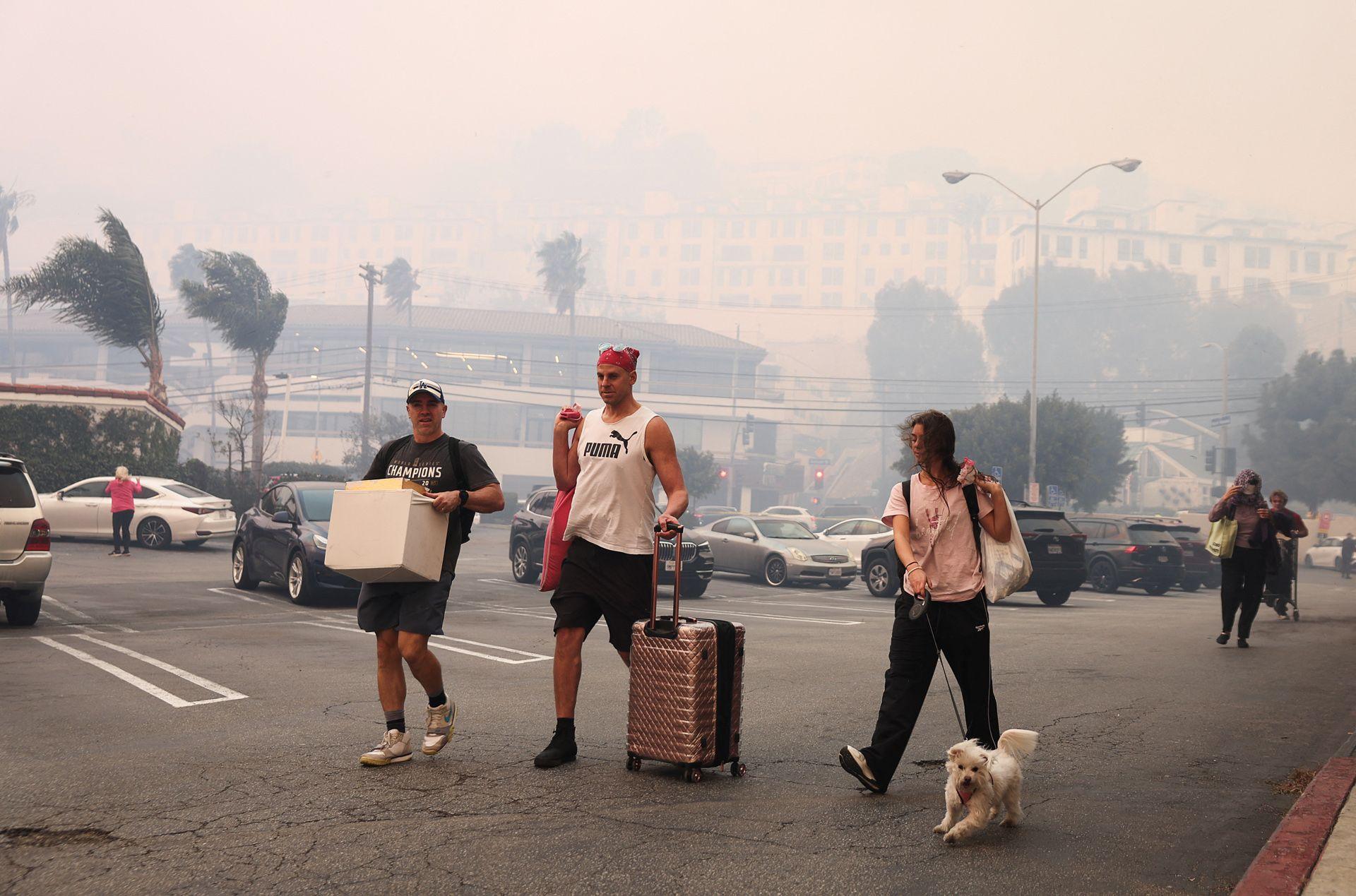 People walk through a car park carrying luggage and belongings, with a smoky background and palm trees bending in high winds, on the west side of LA.