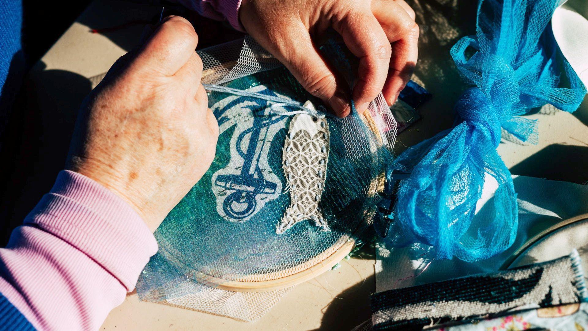 A close up image of a women's hands while she is sewing. The edge of her pink long sleeved top can be seen. She is working on an embroidery which shows a cut out of a fish and a cut out of a blue anchor on blue sparkle fabric. There is a blue tulle fabric bow nearby as she is at a sewing workstation.