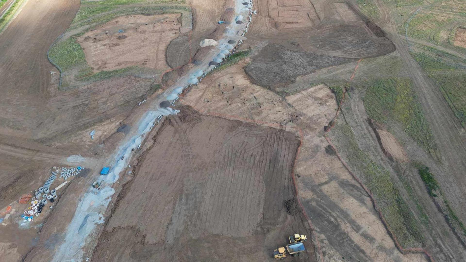 High aerial view of a building site, showing churned up ground, some grassed area, and pipes and other construction equipment and a two tipper trucks