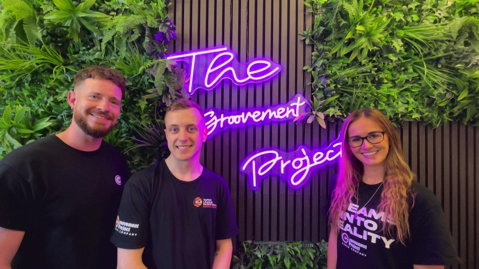 A picture of two men and a woman in front of a purple neon sign. The sign reads "The Groovement Project".  They are all wearing black t-shirts.