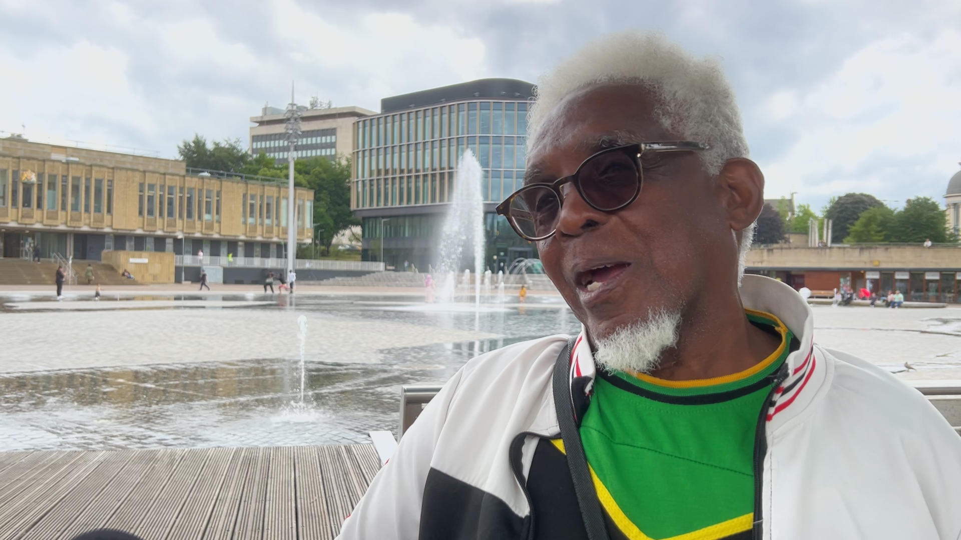 A man with white hair and a beard with sunglasses speaking outside in front of the mirror pool in Bradford