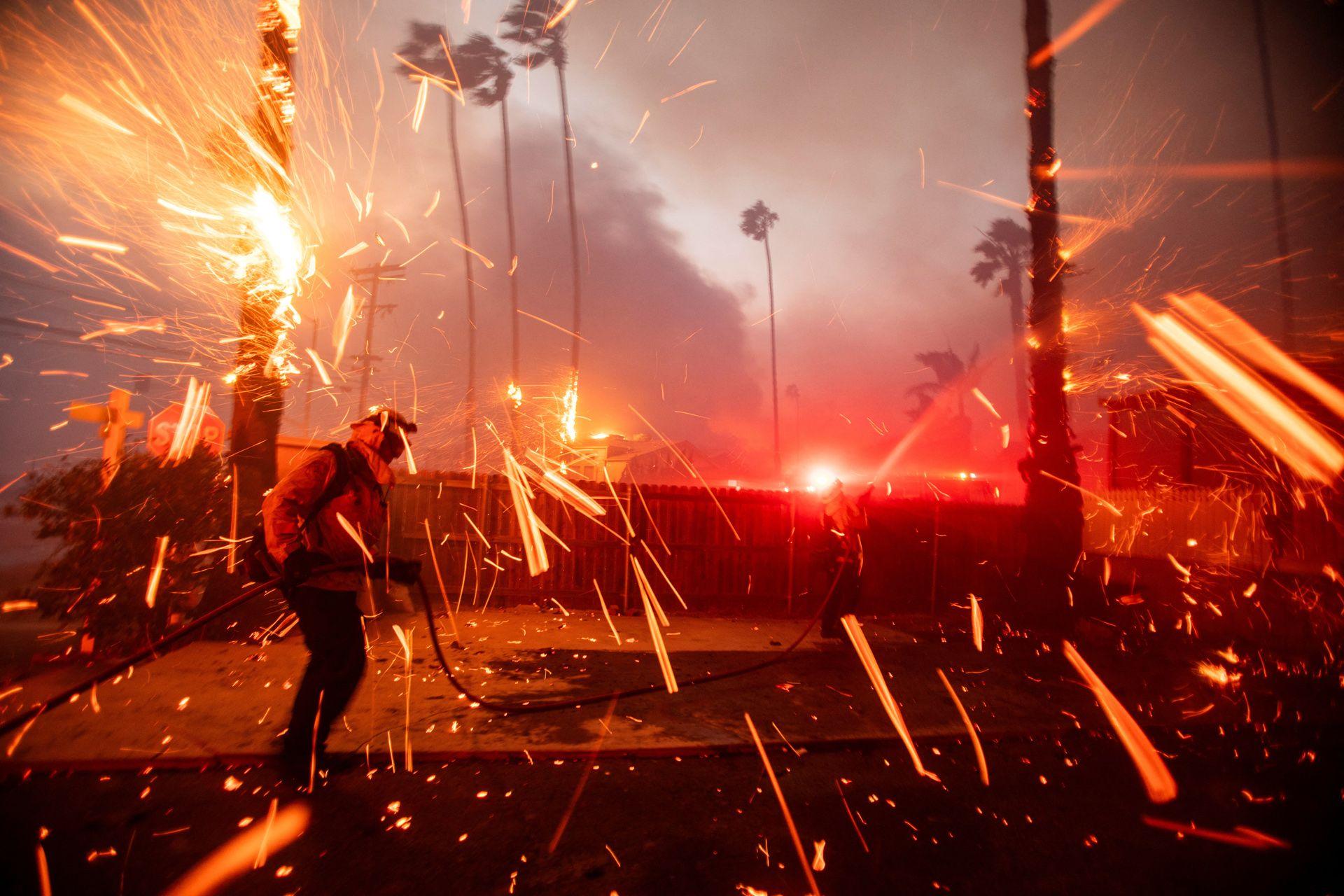 Sparks and debris fly through the air with a firefighter holding a water cannon in the foreground and a fence and trees in the background, on the west side of LA.