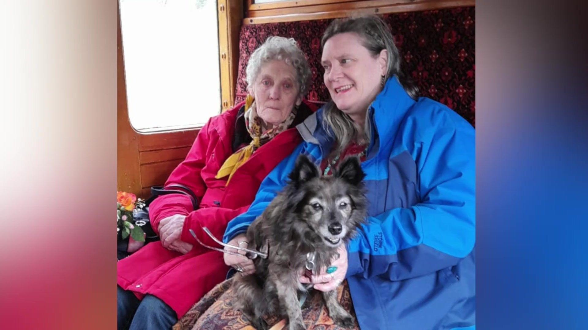 Miriam Souter has short grey hair and is wearing a pink coat and yellow scarf. Sat next to her is her daughter Phazar, who has platted brown hair and is wearing a blue coat. On Phazar's lap is a medium sized black dog.