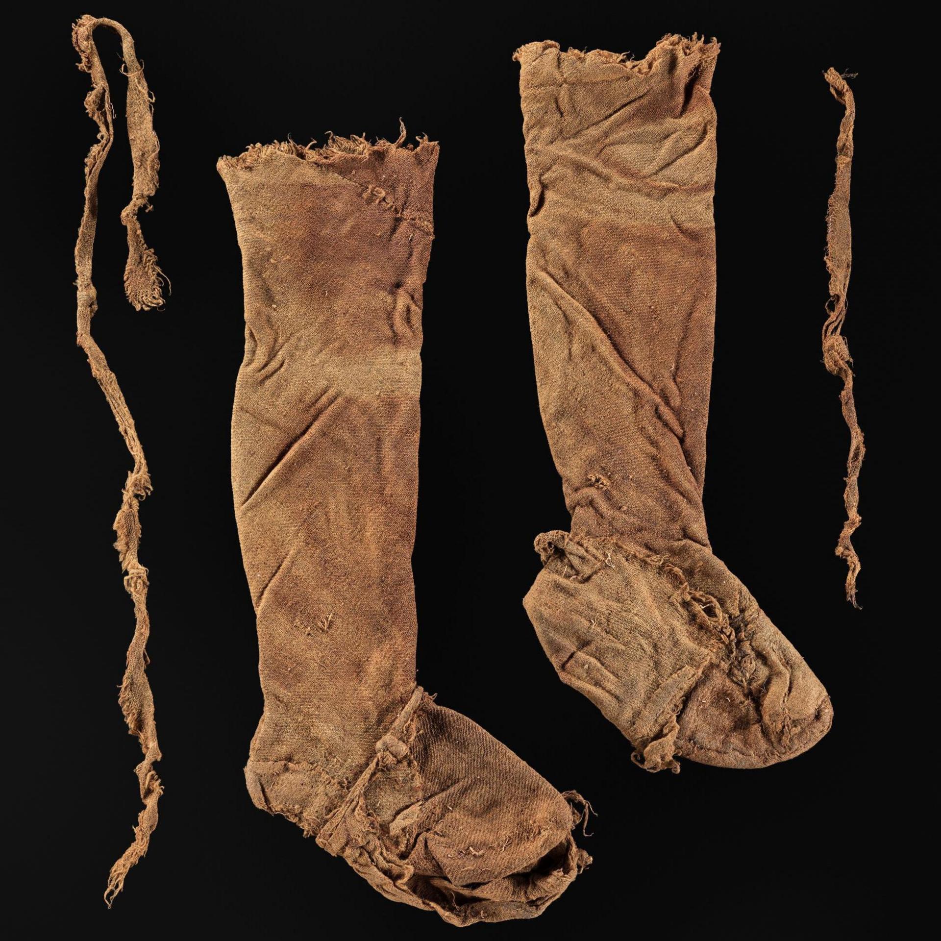 The leggings are long and stained a brown by the peat. The fabric is crumpled. They have been laid next to two long ties on a black background.