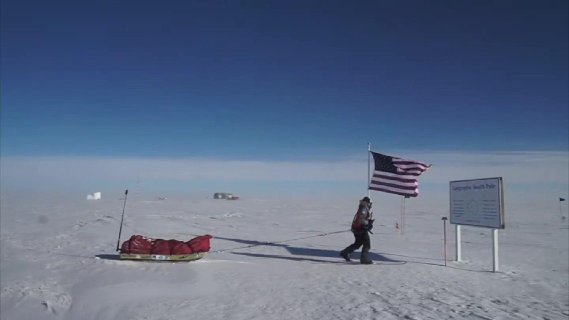 Surrounded by snow Cat Burford skis to a sign making the South Pole. She is dragging a sled with equipment covered by a red material, with the US Stars and Stripes flag just behind her.