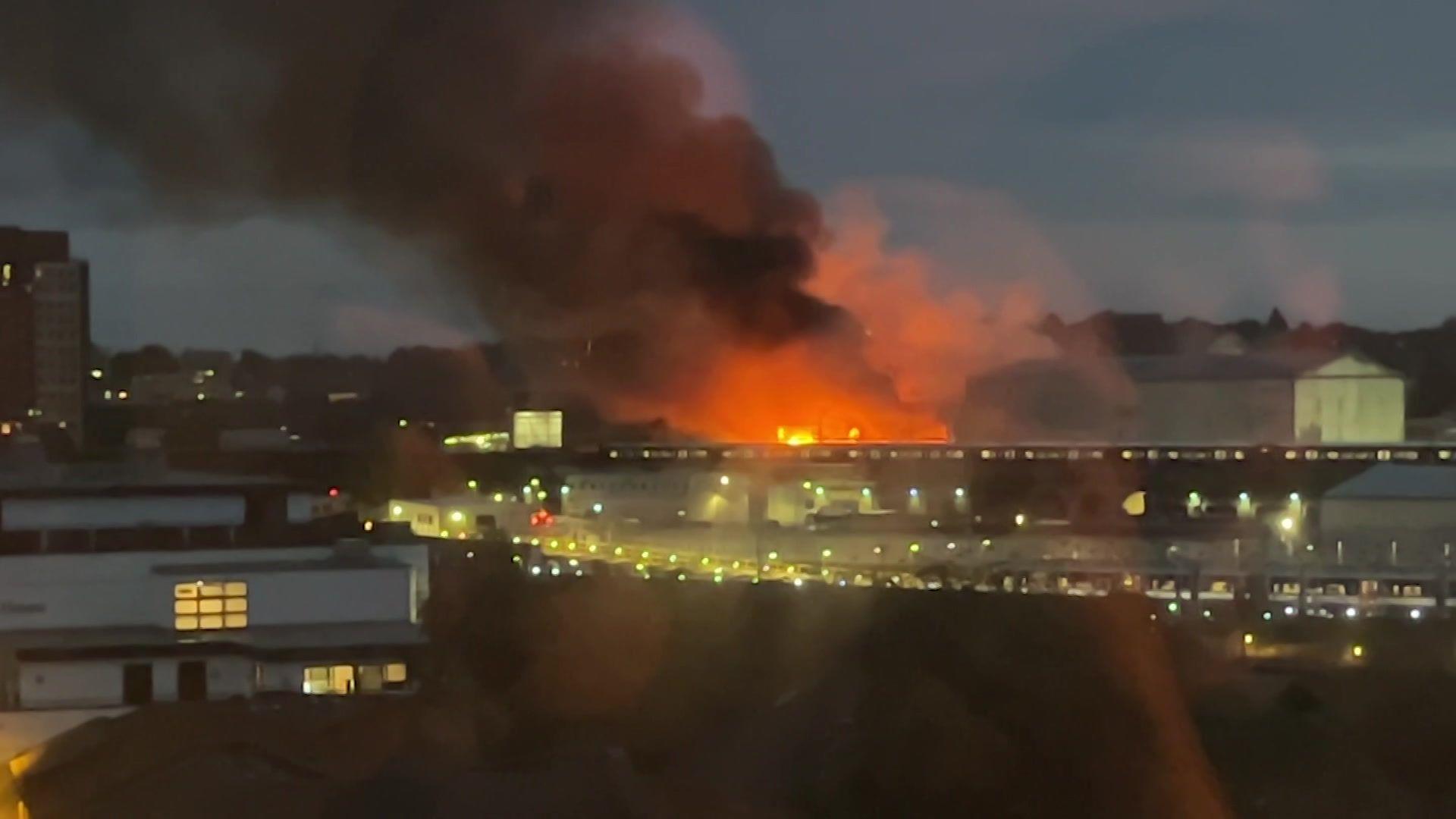 An elevated night view shows a distant fire glowing bright orange with dark plume of smoke rising over industrial buildings and urban skyline.