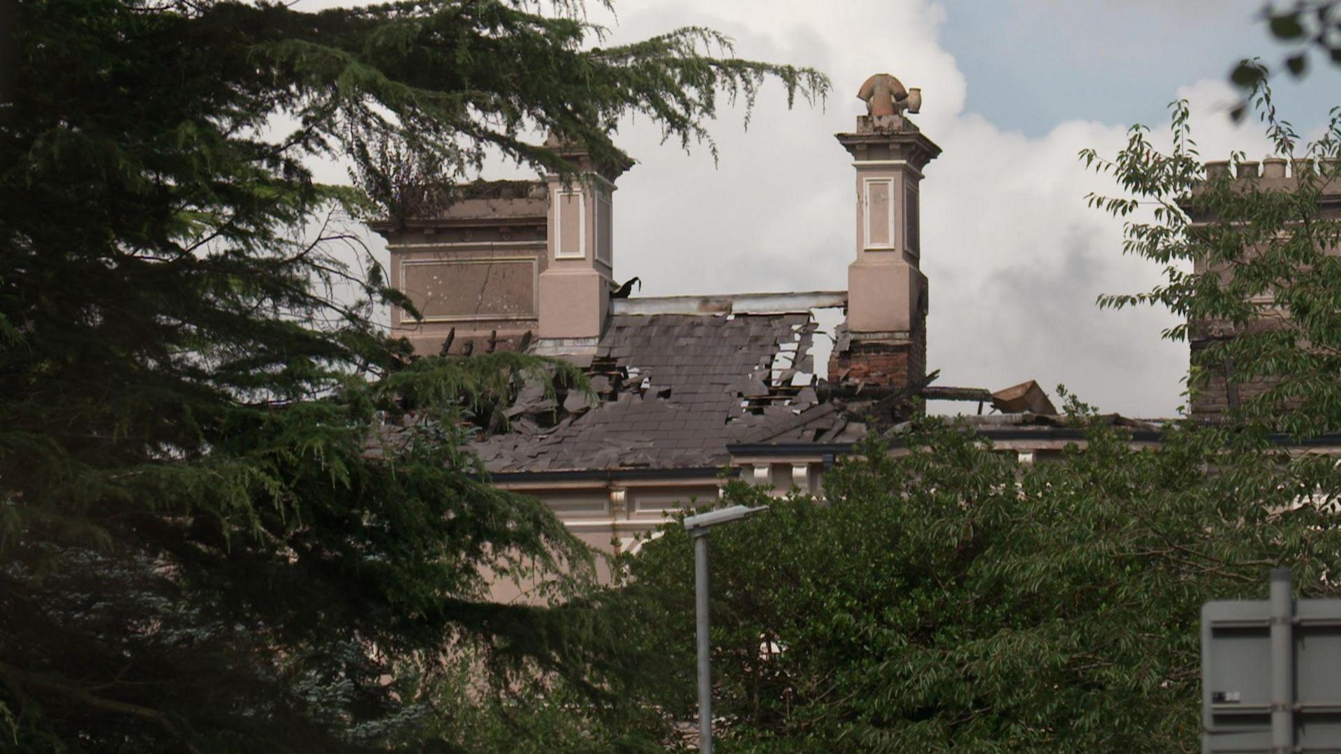 A close up of the fire-damaged roof of Duncreggan House. Large parts of the tiled structure are completely missing. Large ornate chimneys are protruding from the roof. The view is partially obscured by trees.