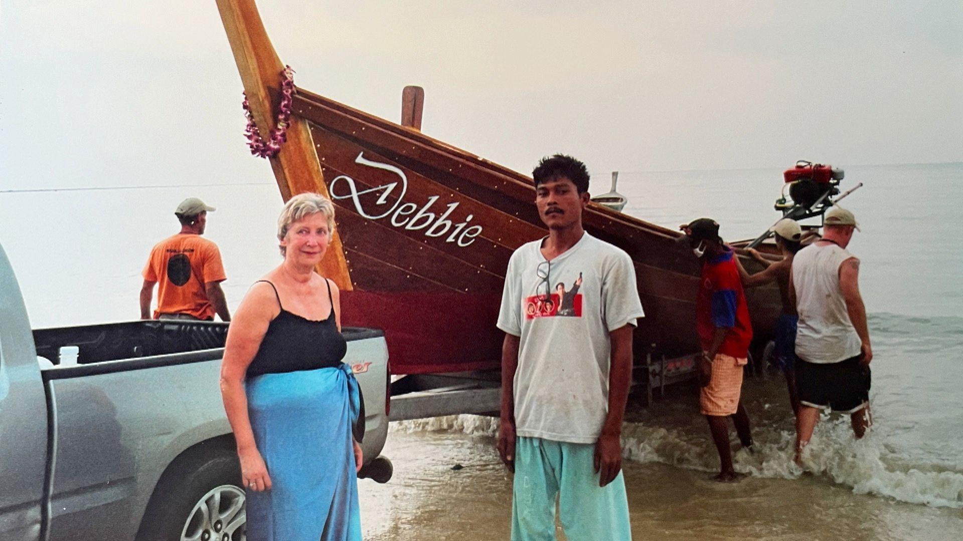 Margaret Garlick standing next to a Thai fisherman on the beach. Behind them is a boat with the name Debbie inscribed on it.