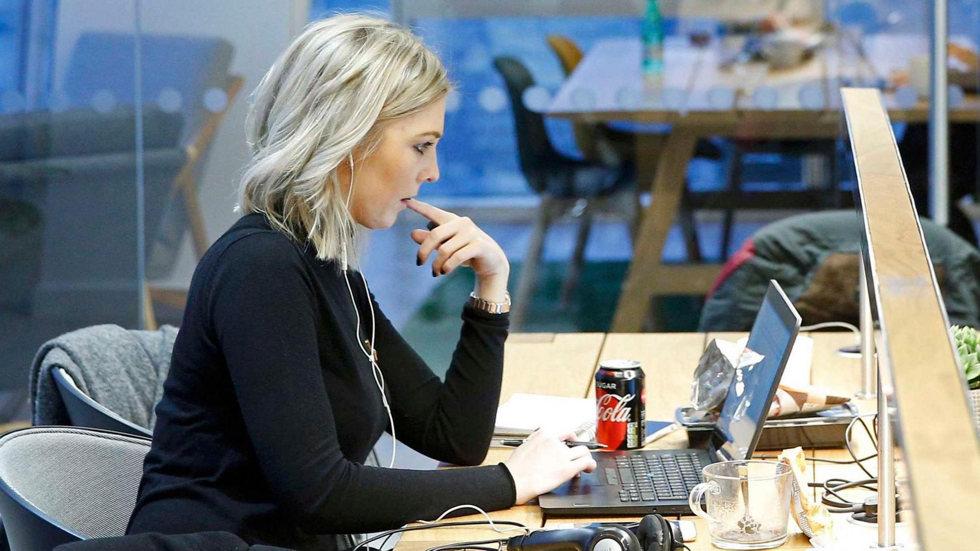 Woman with blonde hair sitting at a desk in front of a laptop in a well- lit office. She is wearing a black top and a pair of white in-ear headphones in her ears. She is holding her left index finger in her mouth while using the trackpad on the laptop keyboard.