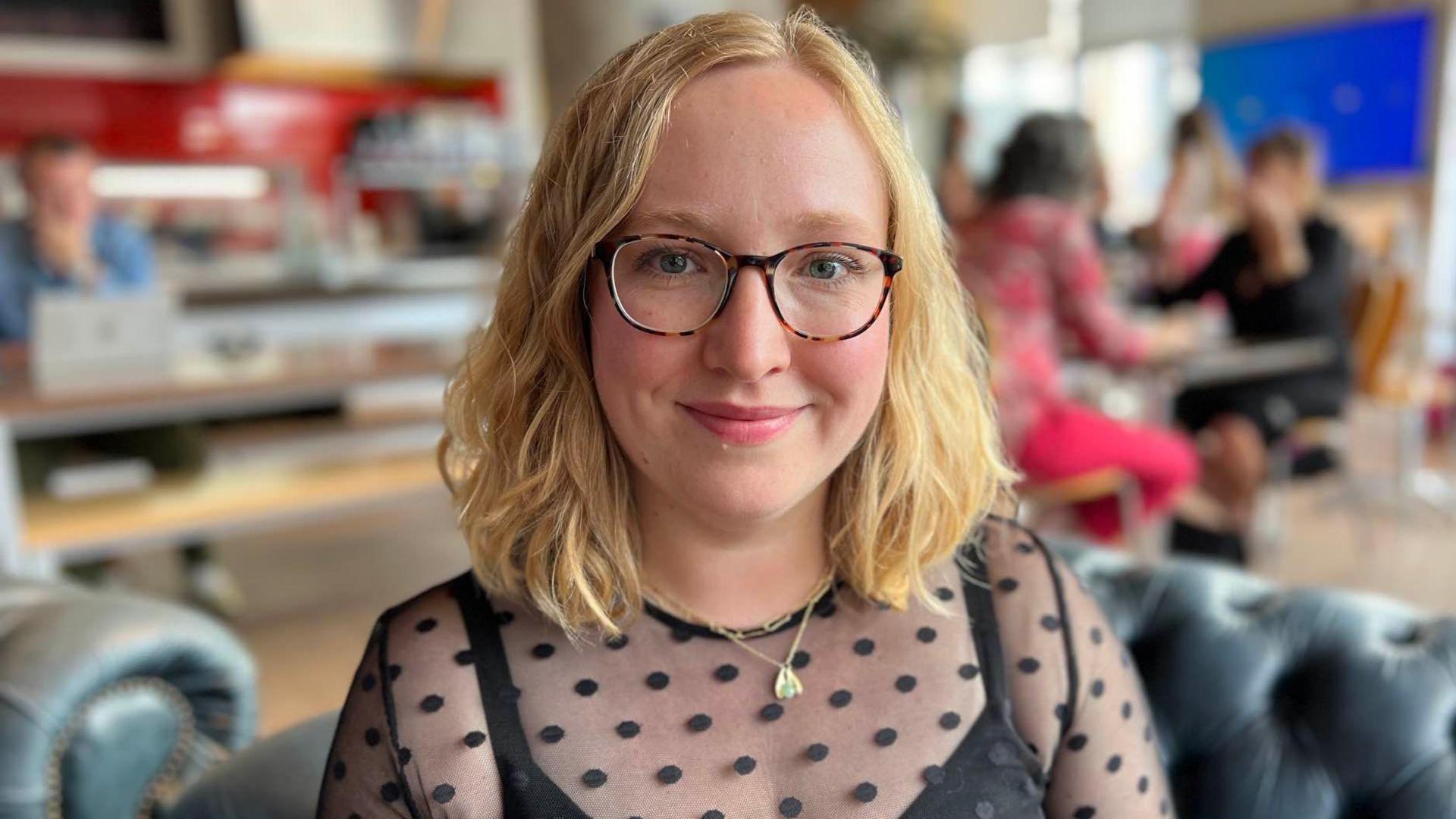 Bryony Hope with blonde hair and dark framed glasses, wearing a black top with black spots on net, smiling towards the camera, sitting in a cafe