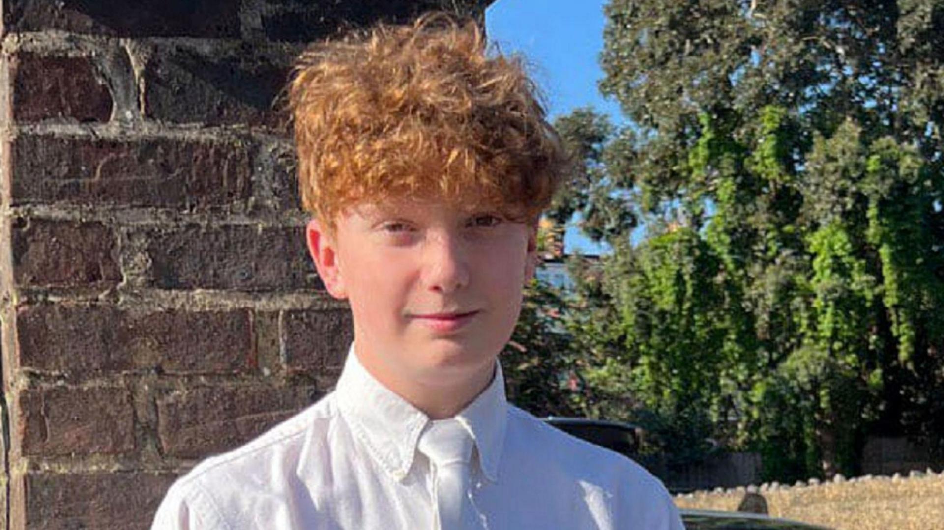 File image of Harry Pitman, a boy with short curly ginger hair, wearing a white shirt and white tie while stood in front of a brick wall and a tree