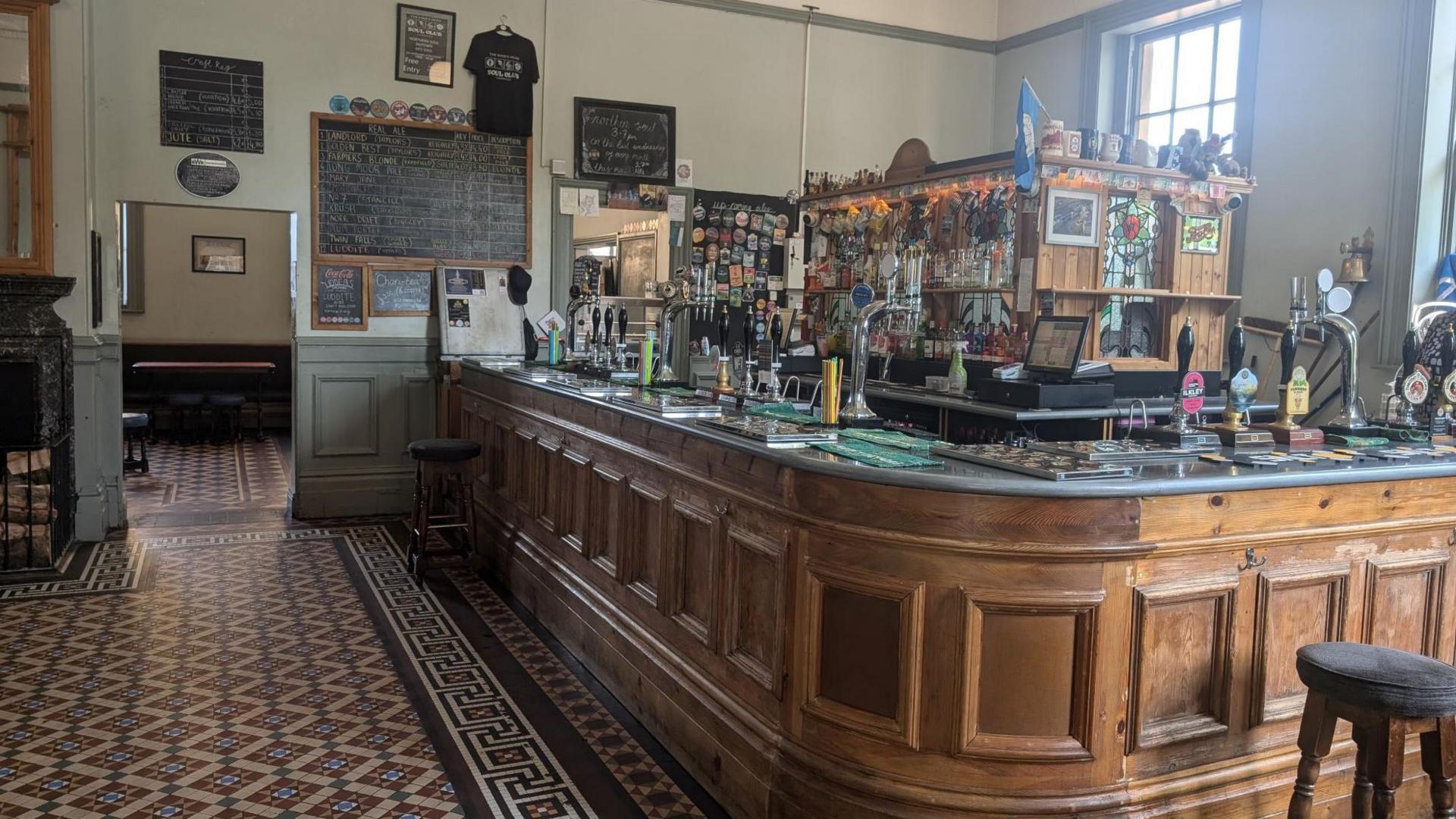 The inside of a pub with a Victorian-style tiled floor. There is a wooden panelled bar with several beer pumps and a bar stool in front.