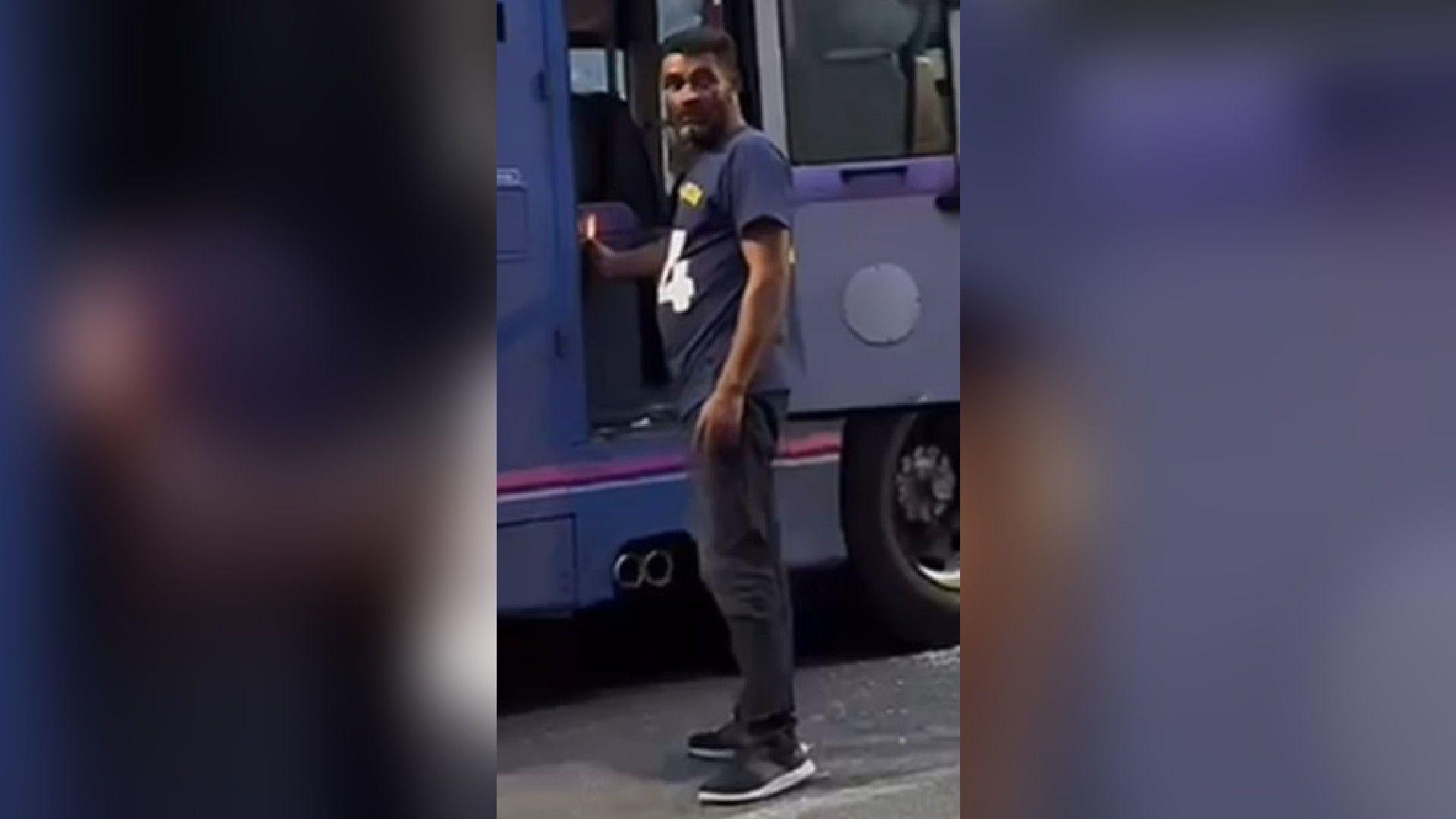 A man in dark trousers and a blue t-shirt holds a lit flame to the back seat of a bus