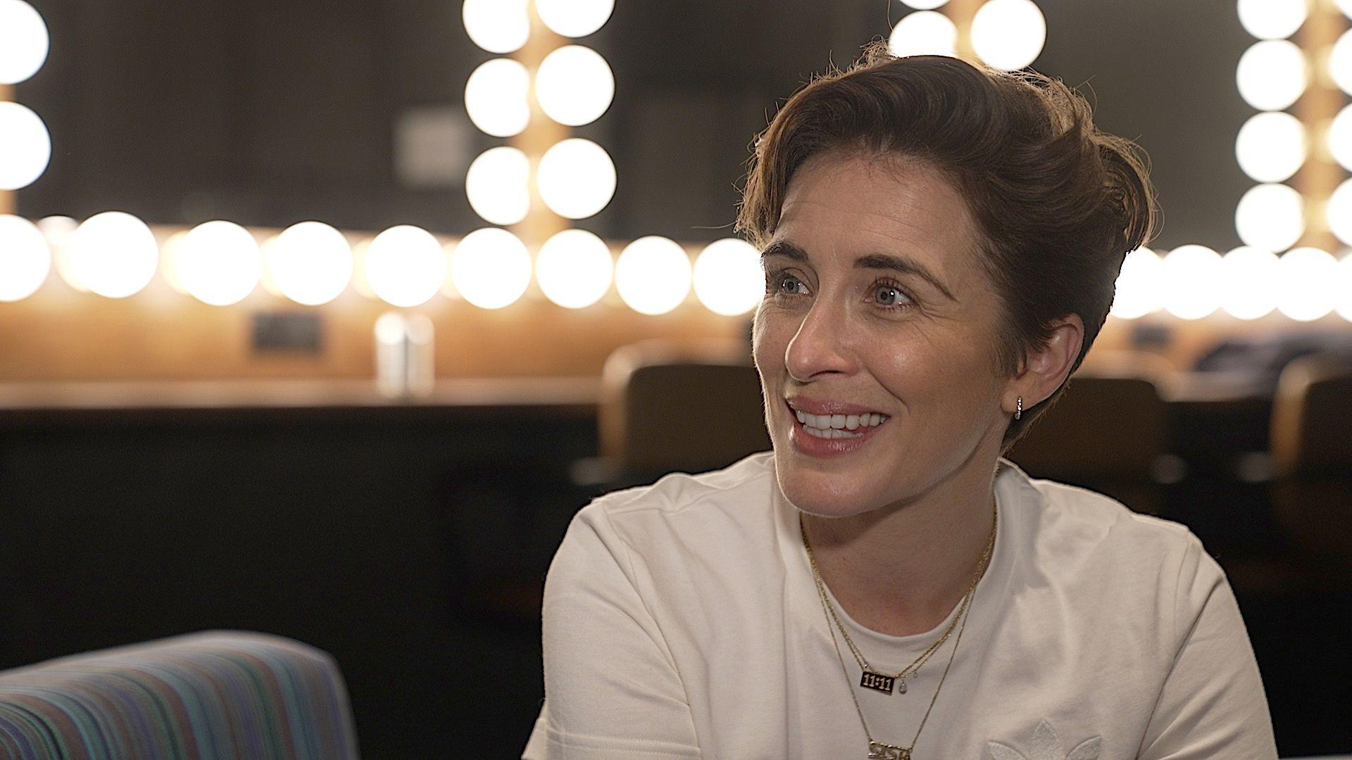 Vicky McClure is seen from the chest up and smiling in what appears to be a dressing room. She is wearing a white top and there are mirrors with lights surrounding them in the background.