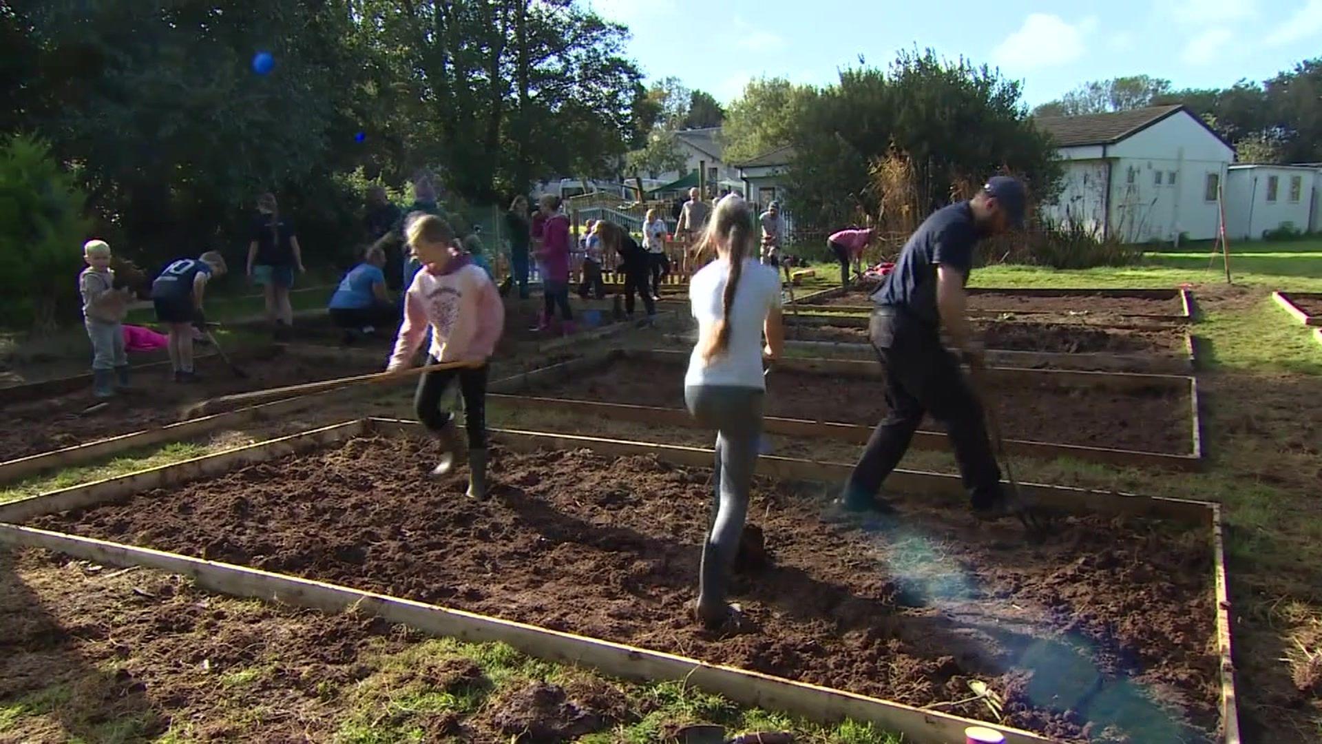 Children stand in a raised vegetable patch wielding garden tools, in the background more patches and children can be seen standing in the soil of the green space. 