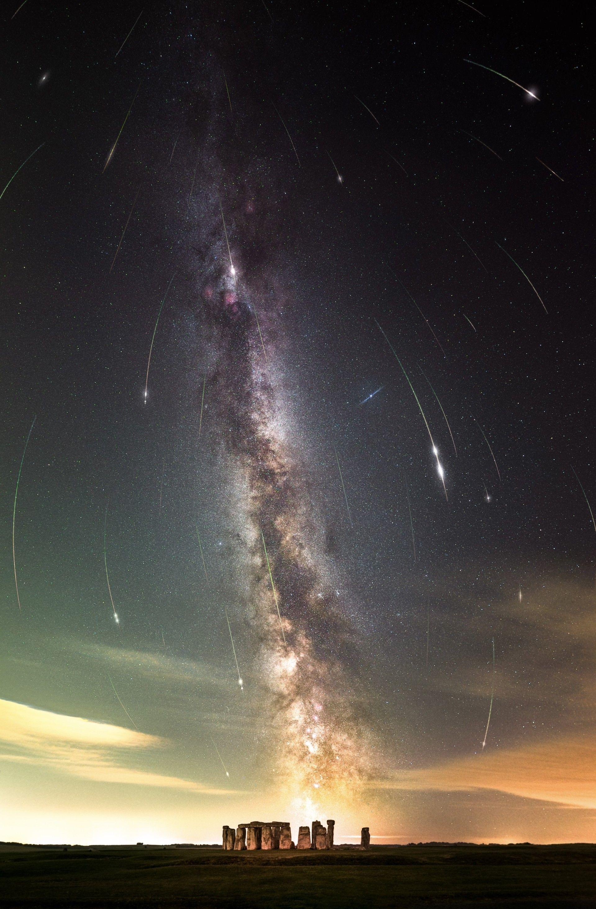 A vertical image - Stonehenge as seen from afar, with the base of the stones lit up slightly. Above the monument, you can see a large vertical cloud shape of white and purple, which is the Milky Way galaxy. Either side of the galaxy, thousands of stars can be seen, and meteors can be seen streaking towards the ground.