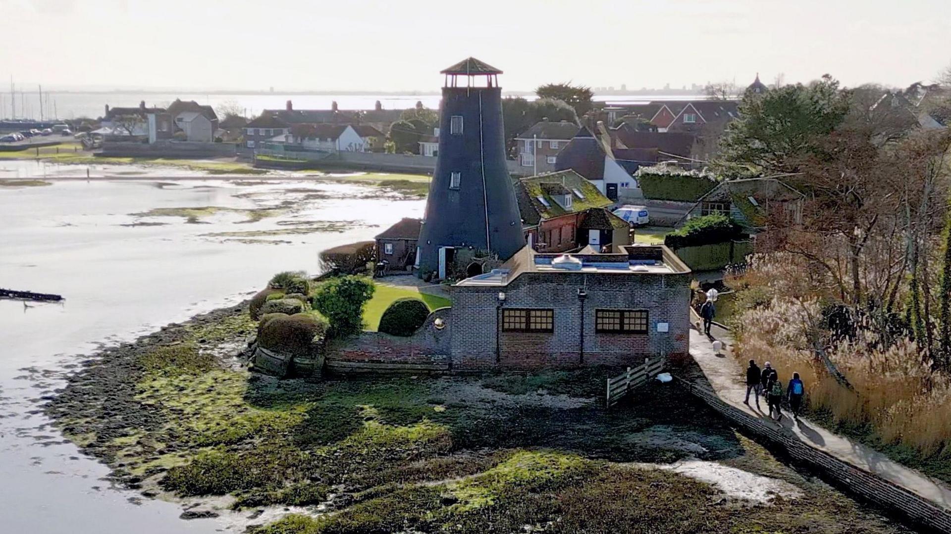 A wide, aerial shot of a part of King Charles III England Coast Path in Hampshire shows four people walking on a path by sea shoreline on a sunny day. The stretch of land between the sea and the path looks muddy and is covered in green moss and low grass. On the other side of the path, there is brown grassland with a few trees with a green field in the background. In the middle of the photo, there is a stone building and a round tower.