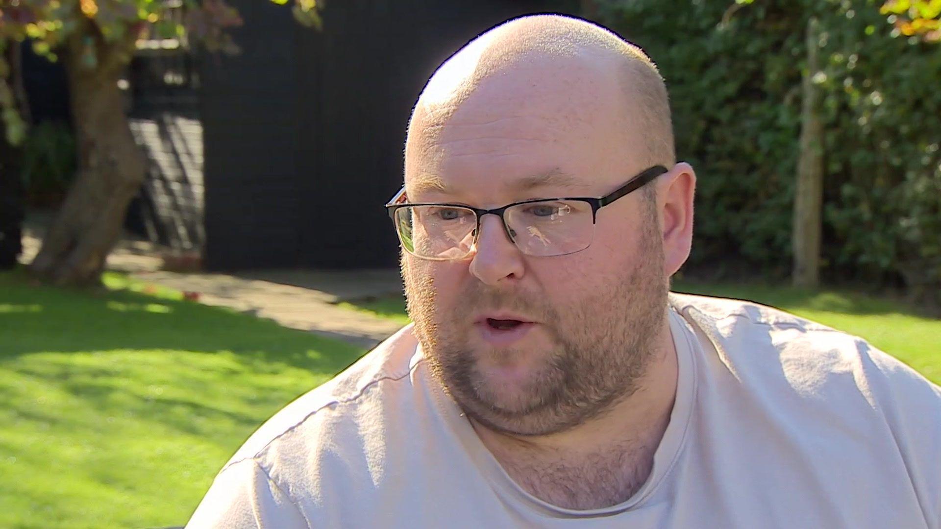 A man wearing a light pink t-shirt and glasses. He is sitting in a garden.