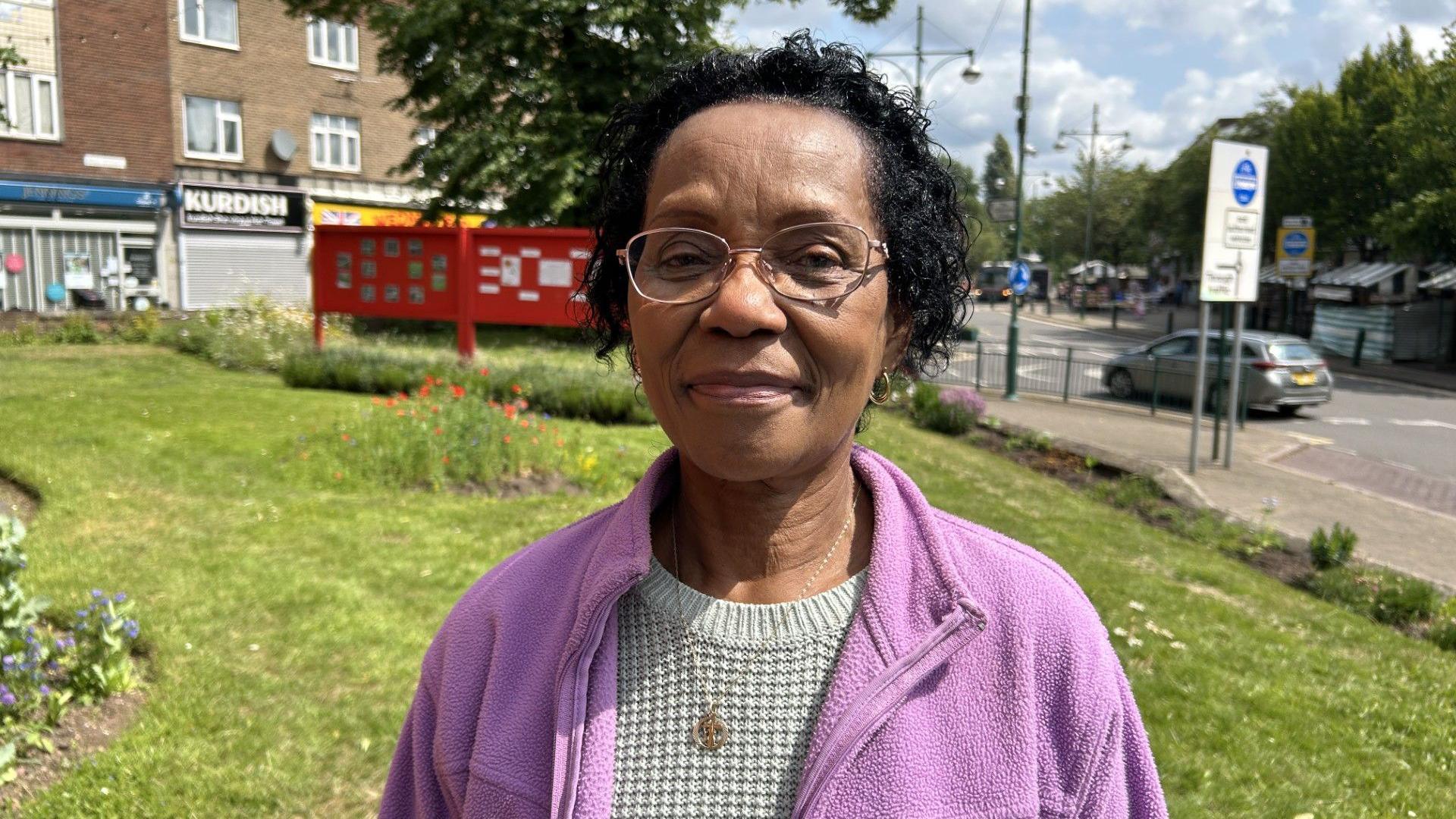 Norma Thompson in Wednesfield town centre. We can see some shops and cars behind her. She has short black hair and she is wearing a purple fleece and a grey jumper. She has a necklace on and is wearing glasses.