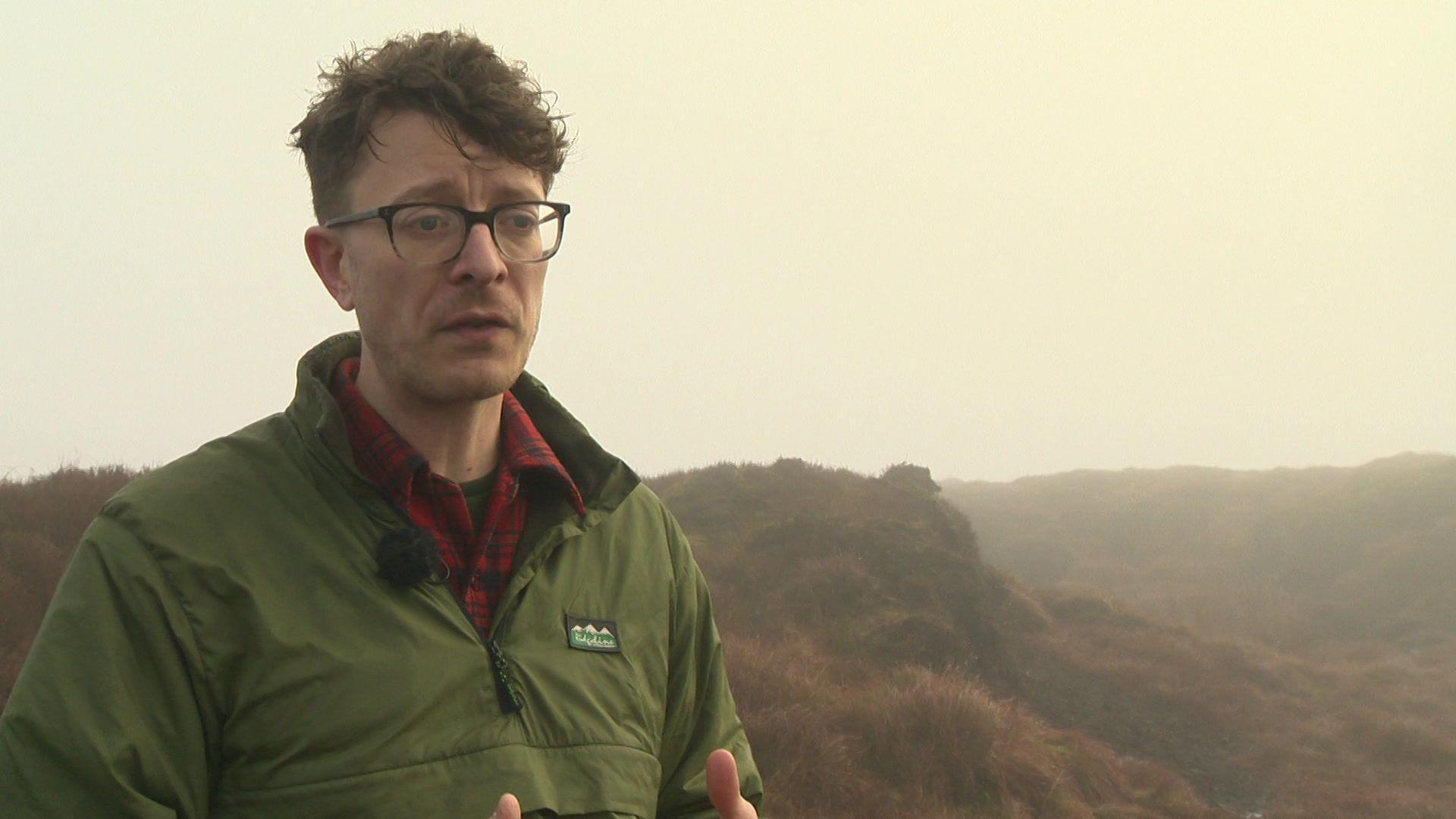 A man with brown hair and black rimmed glasses, wears a green jacket and stands on a hill in the Peak District Moors