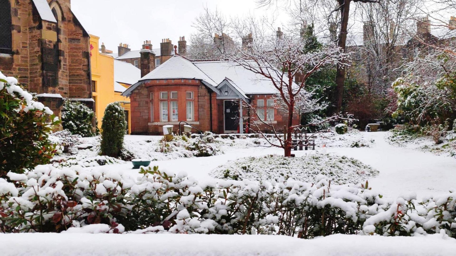 A house covered in snow. The house is made of red brick and has a large ground floor bay window. The roof is covered with snow, as is the garden in front of it. Some greenery from bushes can be seen in the foreground. A reddish-brown tree, which is bare, has snow on its branches. On the left of the image, the corner of a church, made of light and dark stone, can be seen.