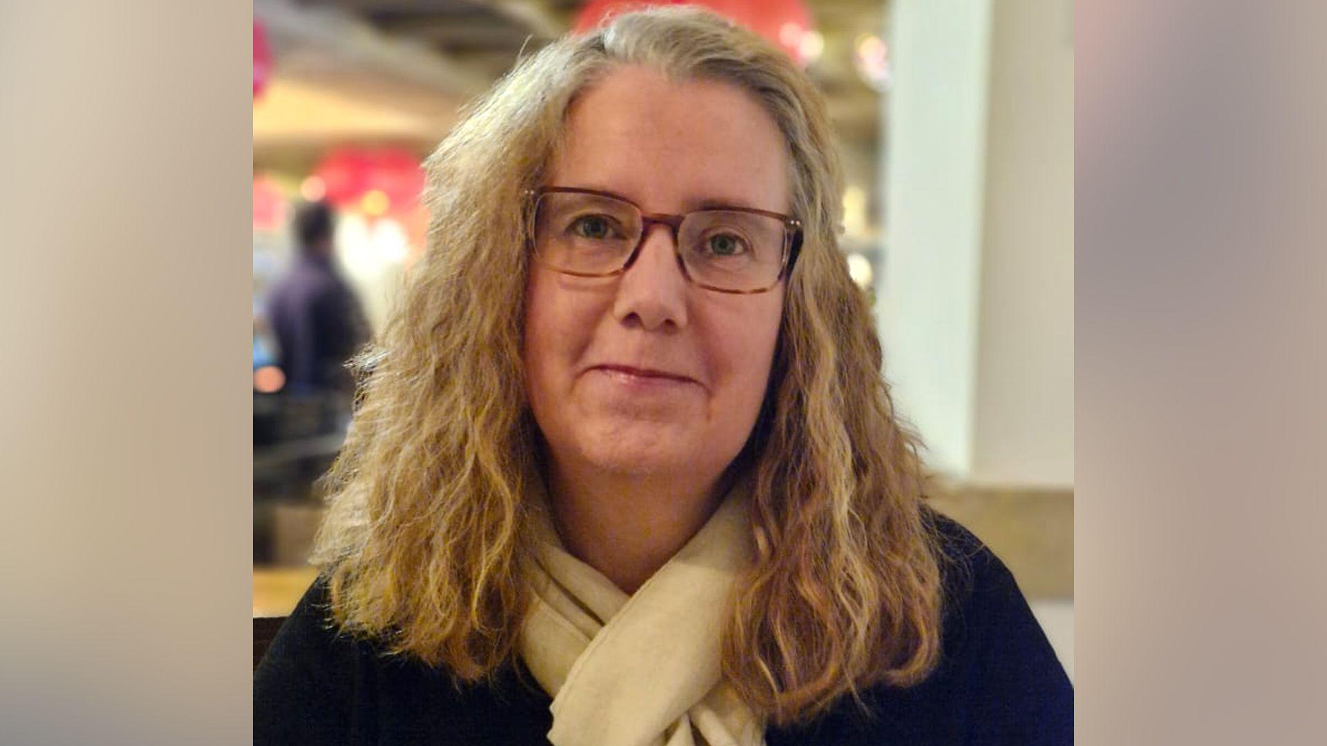 Rachael King, me flokë kaçurrela të çelëta, me syze, një bluzë të zezë dhe një shall ngjyrë krem