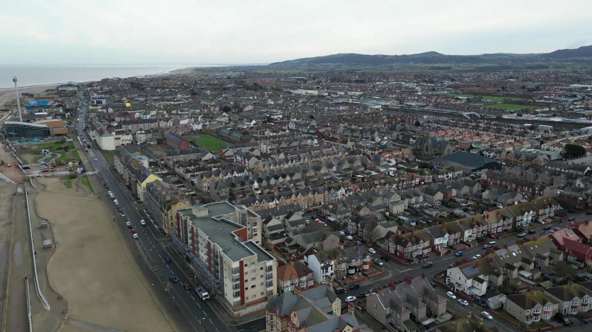 Aerial shot of the west end of Rhyl, with the beach on the left and streets of houses stretching across the shot.  Hills in the far distance.