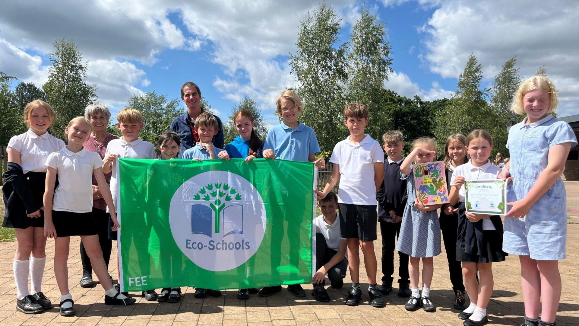 Children in a line smiling holding a green flag that says, 'Eco-Schools'.