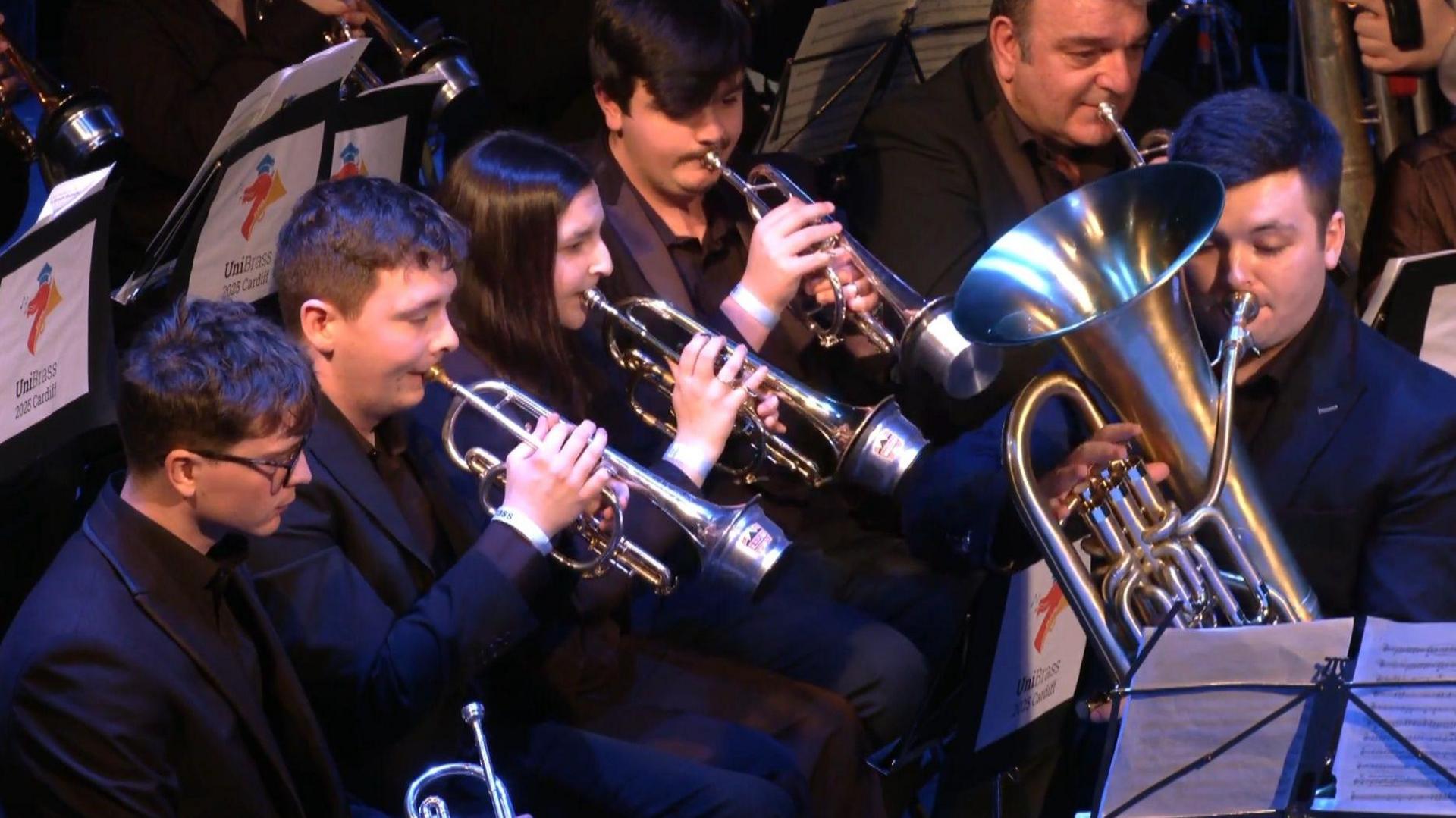 Five men and woman in black suits play brass instruments in a band.