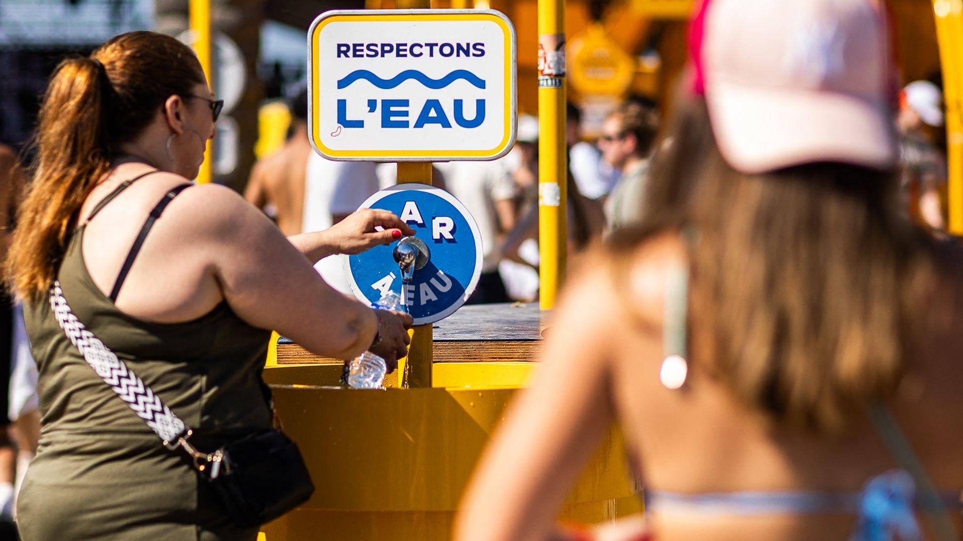 A person with long hair in a ponytail fills a plastic water bottle at a tap with a sign saying 'respectons l'eau' while another reveller waits in line, with people in the background out of focus on a sunny day at Les Deferlantes music festival in Le Barcares on 29 June.
