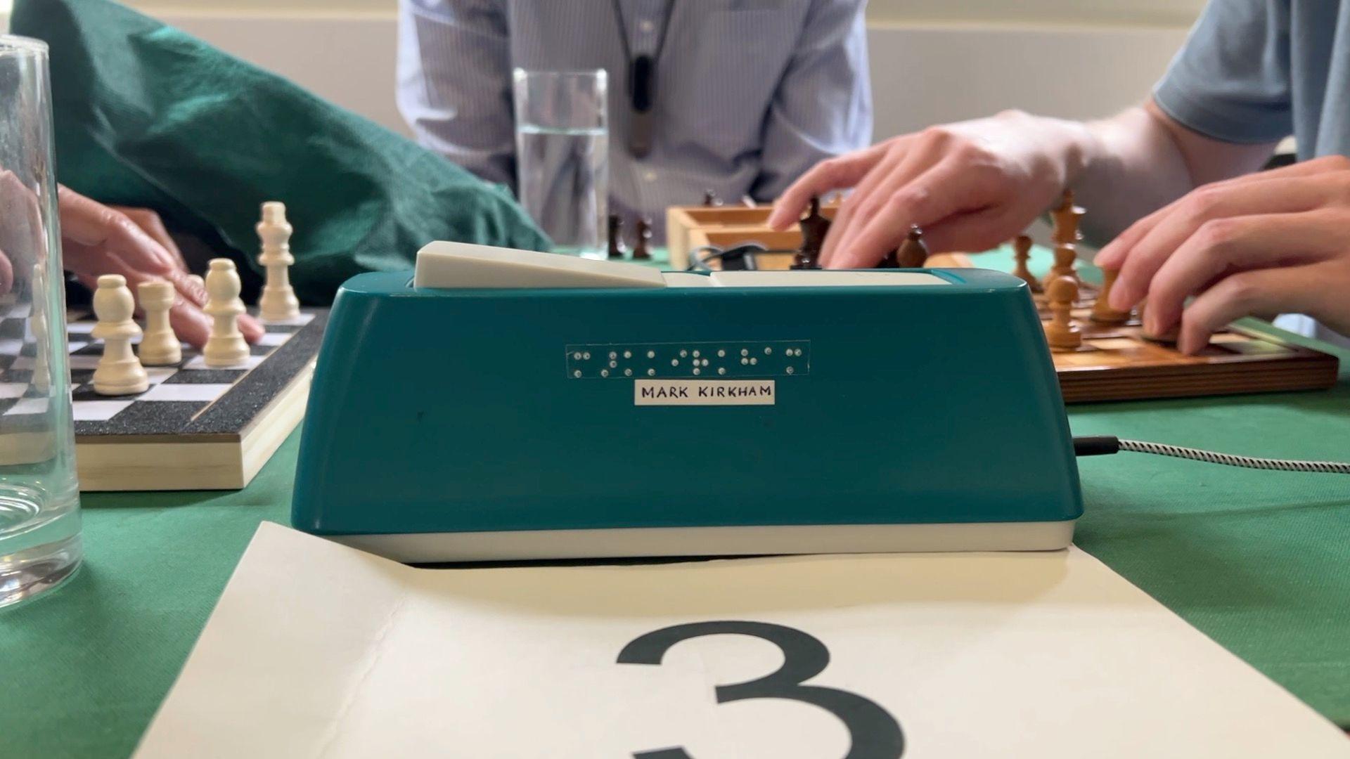 An image of players playing chess at a table with an invigilator in the middle. In the foreground of the image, the table number 3 is shown on a piece of paper, and the clock used to keep time for the game is visible. It has braille printed on it along with the name "Mark Kirkham" on a label. In the background, two boards are visible with hands on them feeling pieces, with an invigilator sat in the middle between them.