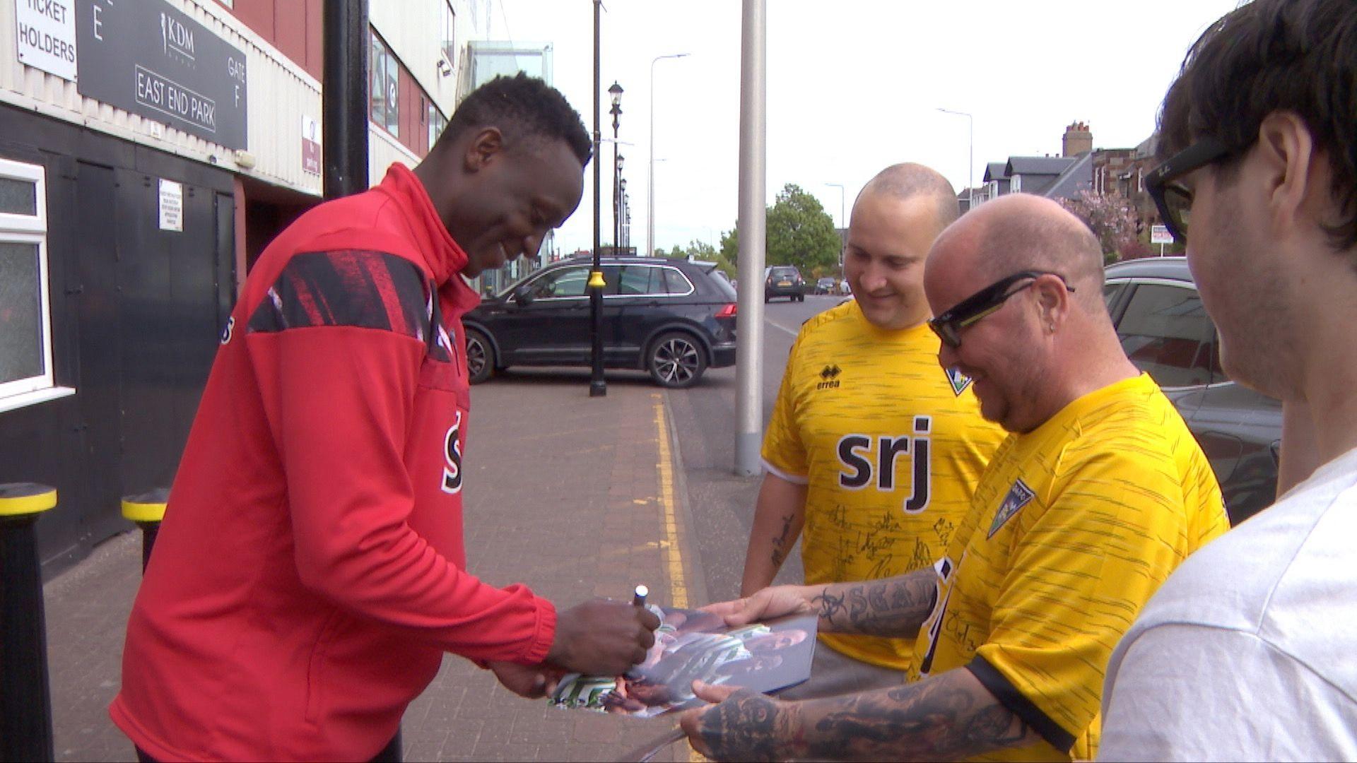 Victor Wanyama wears a red training top and smiles as he signs pictures of himself playing for Celtic for two football fans wearing Yellow Dunfermline Athletic away shirts covered in signatures outside the club's East End Park stadium