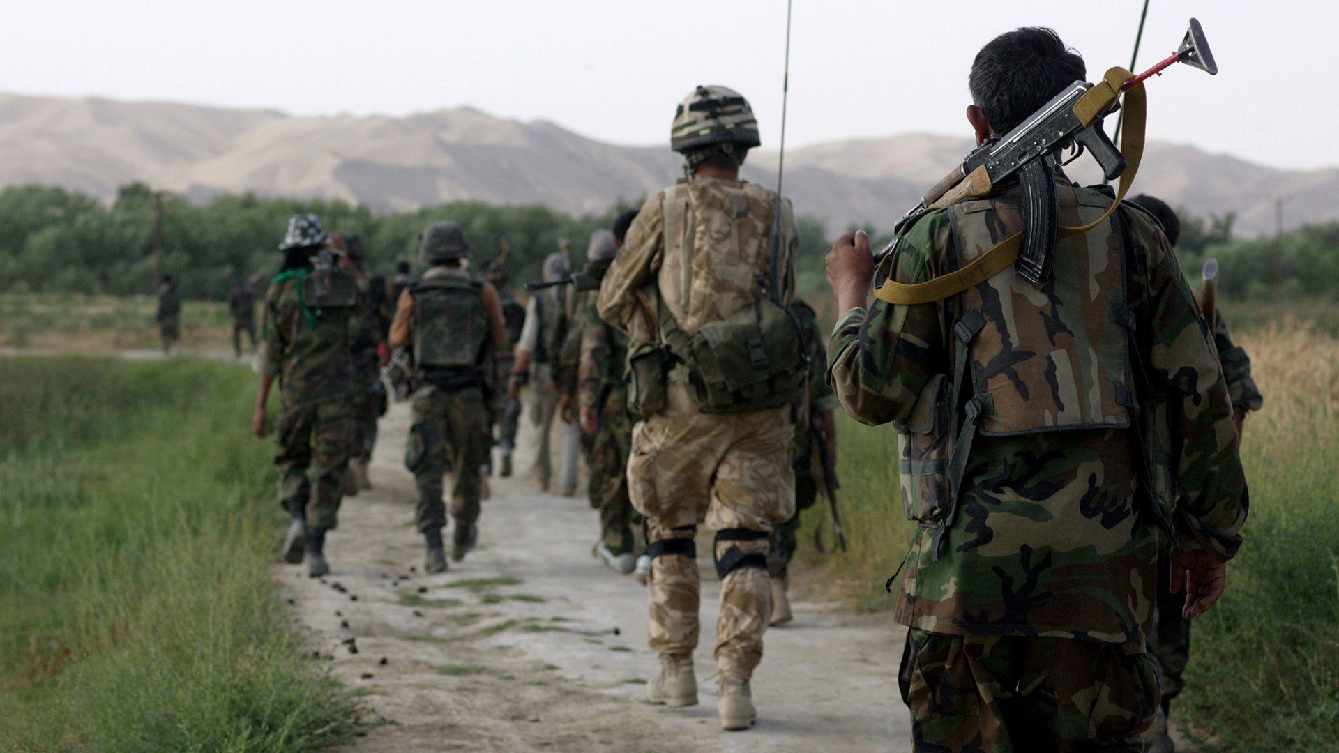British Army soldiers patrol with Afghan National Army soldiers with their backs to the camera, walking down a path through a grass field with mountains in the background, in Helmand Province in 2007.
