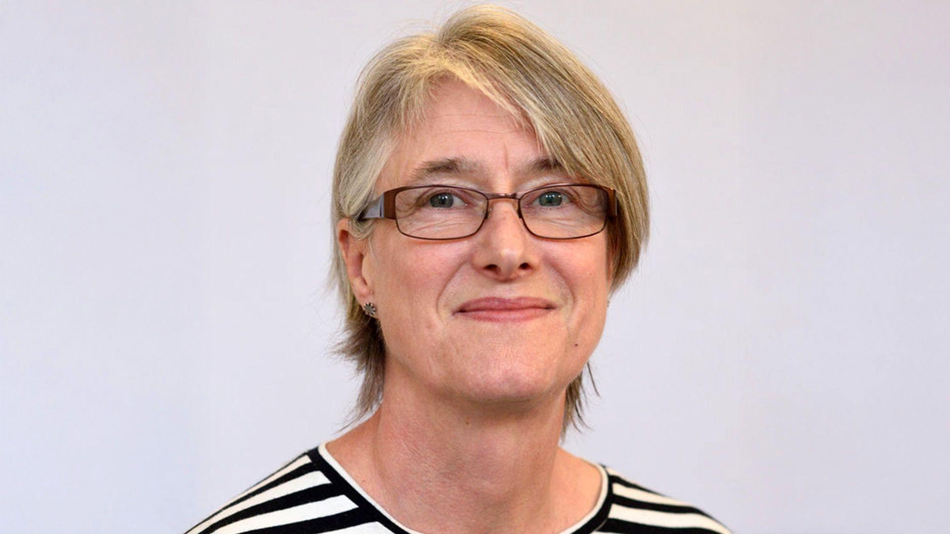 A portrait photo of Prof Hester, who smiles into the camera. She is wearing glasses, a striped top and her hair is cut short.