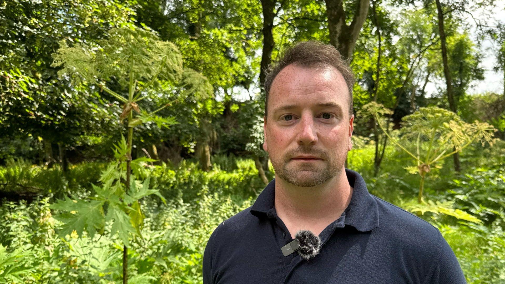 A man with short brown hair is pictured in a wooded area. He's dressed in a blue polo shirt, behind him is a lot of grass and greenery. 