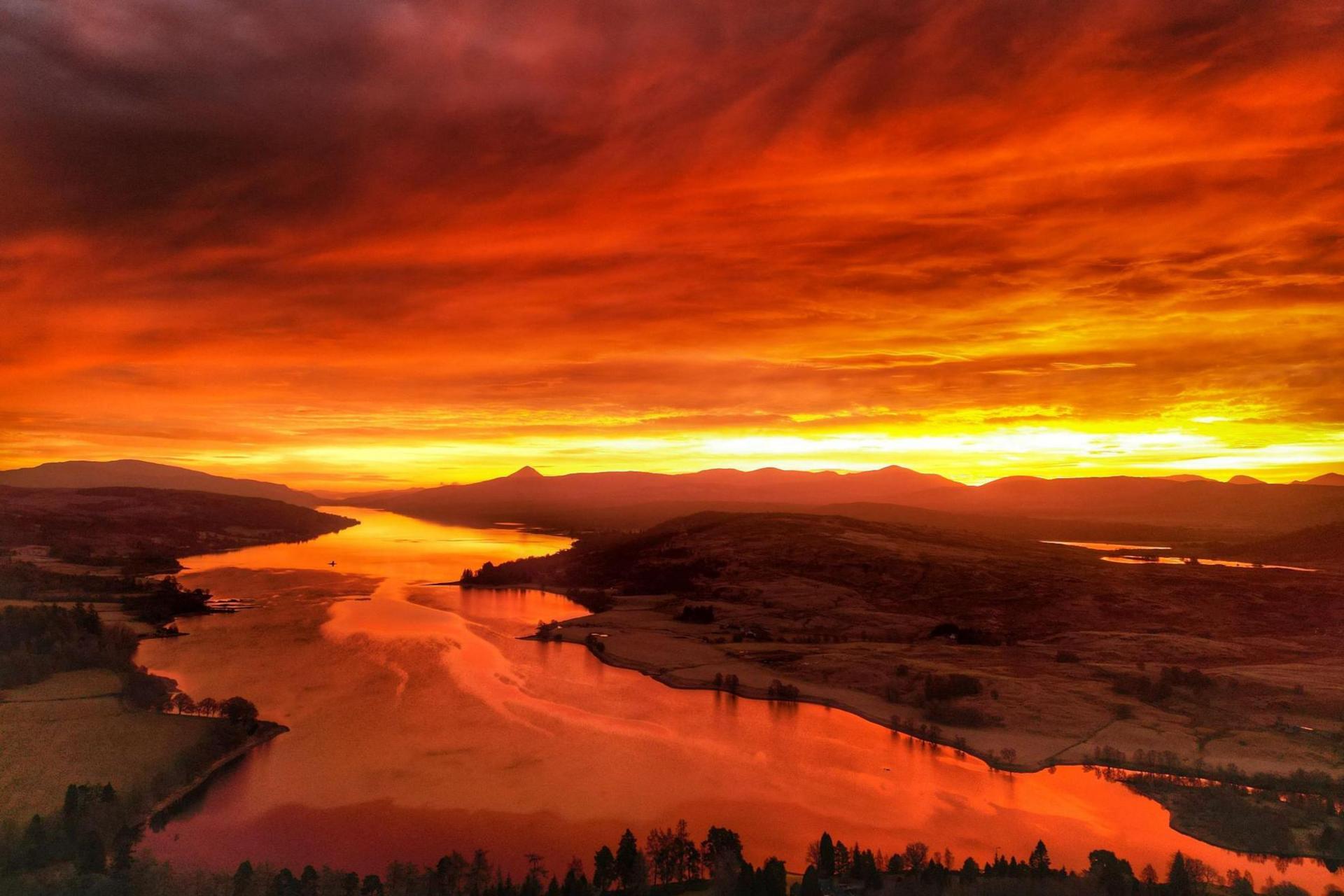 The red sky is reflected in water below. It is a rural landscape of fields and trees.